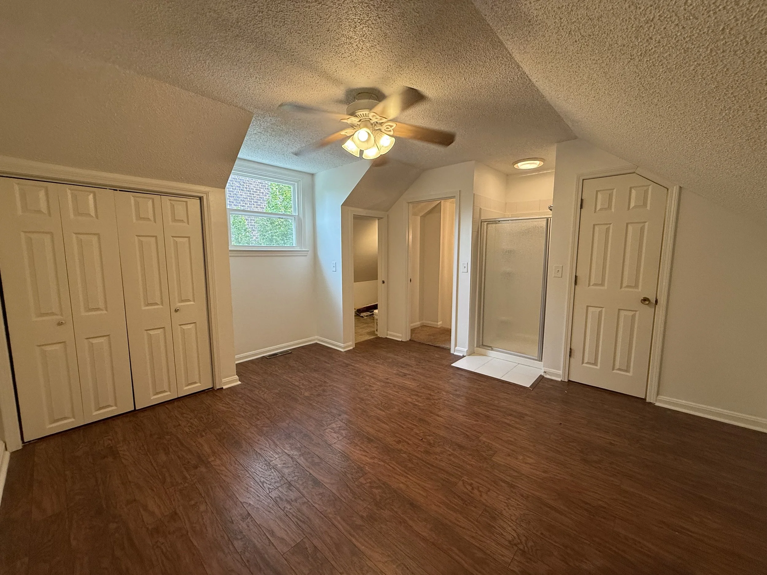 Room with brown wood flooring, white walls, closets, a window letting in natural light, ceiling fan with lights, and an door leading to a bathroom with tile flooring.