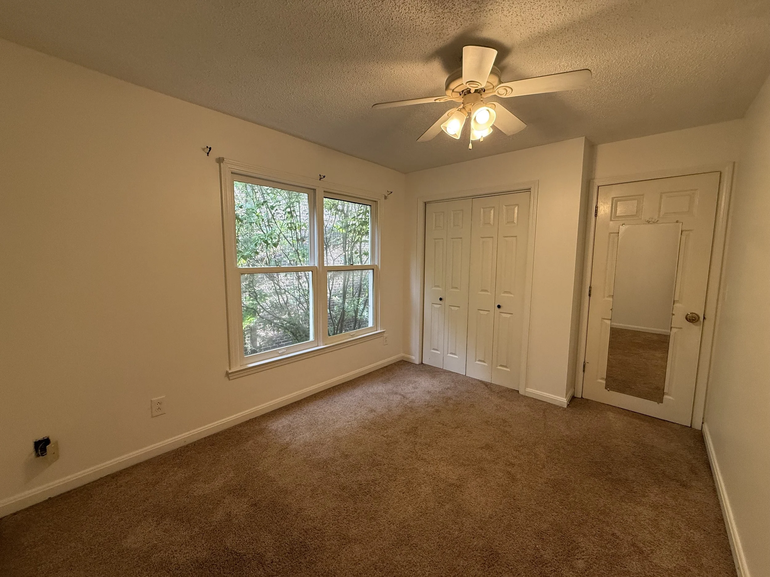 Empty bedroom with beige walls, a large window showing greenery outside, a white closet with double doors, a door to another room, a ceiling fan with lights, and brown carpeting.