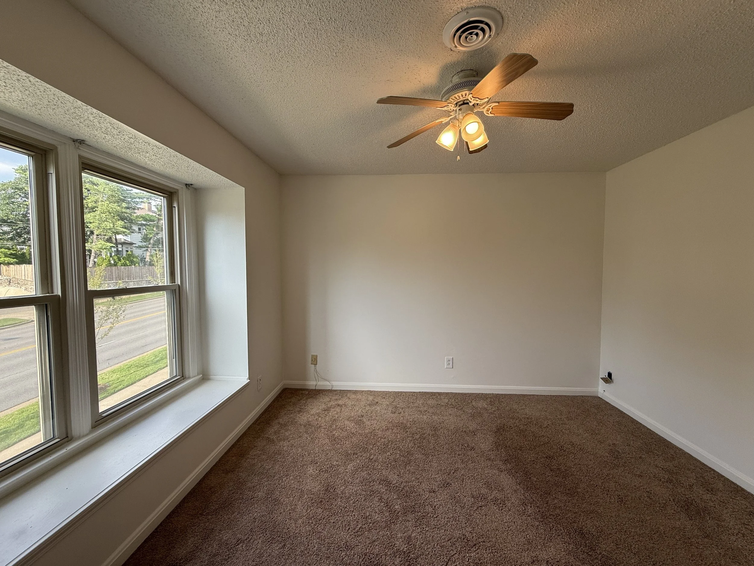 Empty room with large windows, beige carpet, white walls, ceiling fan with lights.