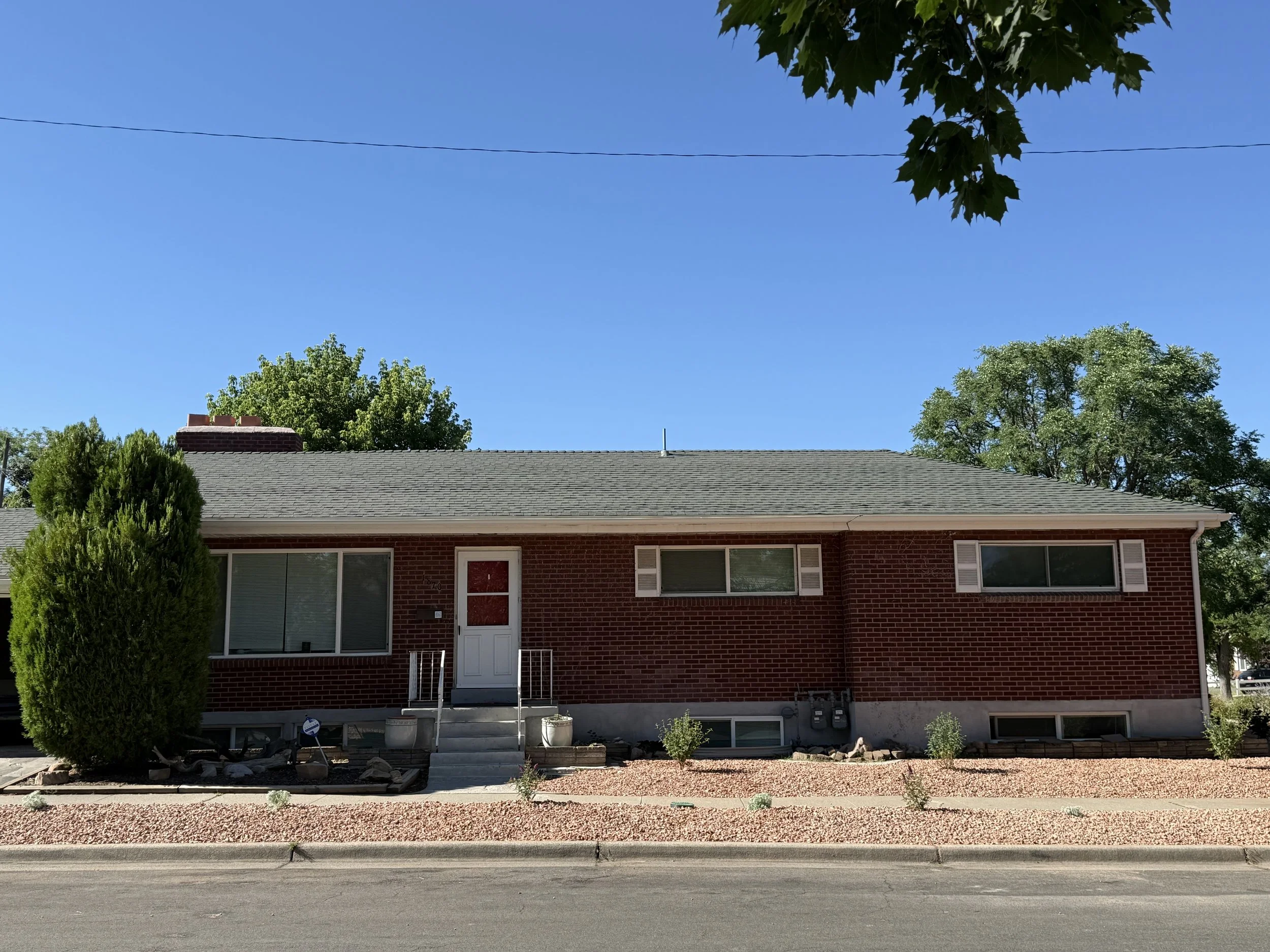 A red brick house with a gray roof, white front door, and a small staircase. Front yard with minimal landscaping, including two small bushes. Tall trees behind the house and a clear blue sky overhead.