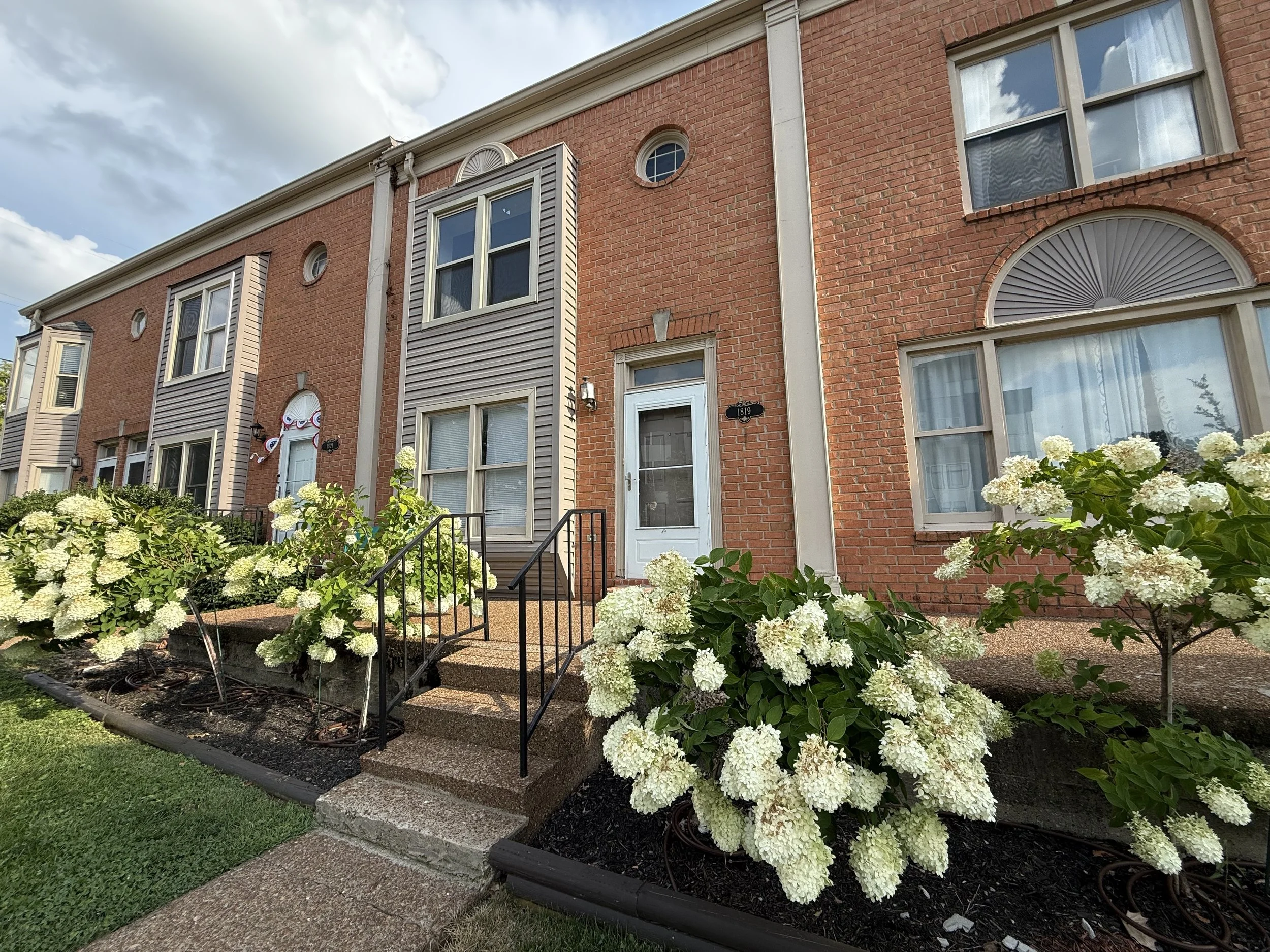 Front view of a brick townhouse with stairs leading to the front door, surrounded by white hydrangea bushes, with cloudy blue sky above.