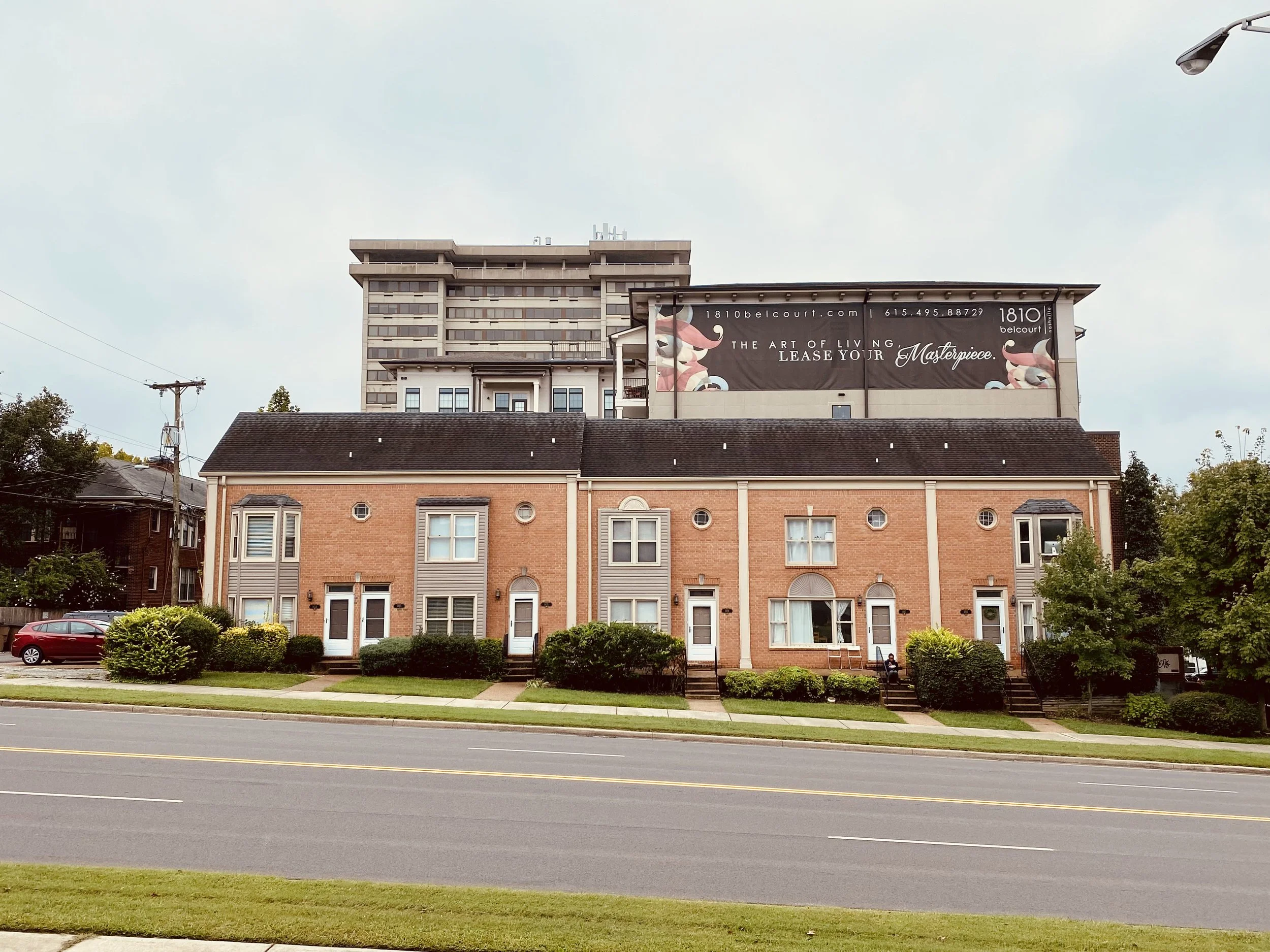 A photo of a street view showing a row of buildings, including a brick residential building with arches, a larger commercial building behind it, and a billboard on the larger building. The street in front has a car, and there are trees and shrubs along the sidewalk.