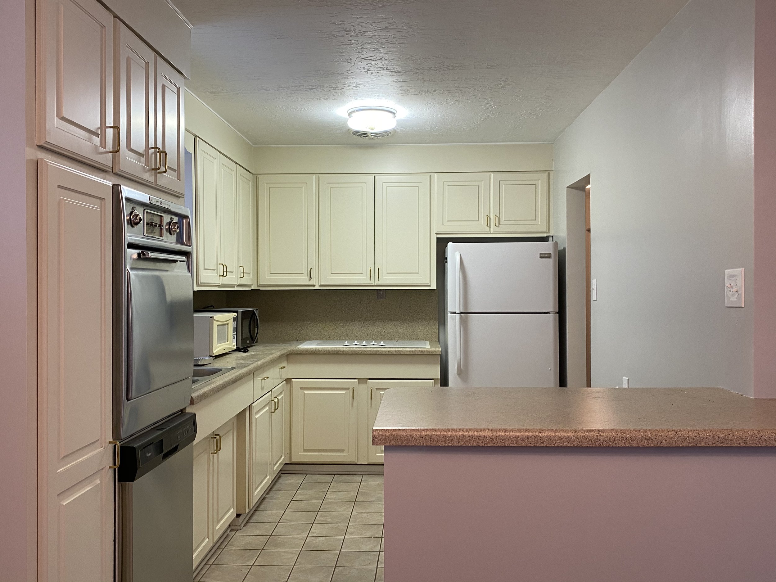 Kitchen with cream-colored cabinets, a white refrigerator, microwave, and a cooktop, with beige countertops and tiled flooring.