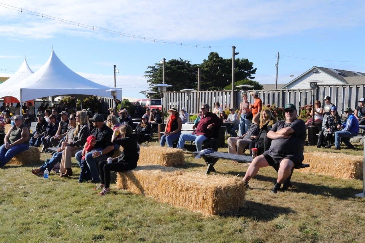 An outdoor event with people seated on hay bales and benches under a large white canopy, with a clear sky and houses in the background.