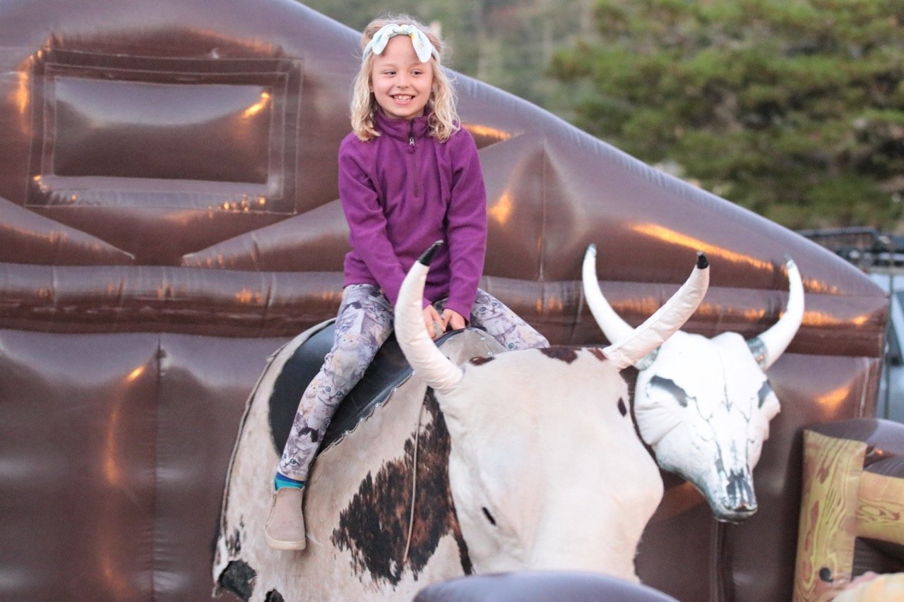 A young girl with blonde hair, wearing a purple jacket and patterned leggings, is riding on a mechanical bull that is shaped like a cow with white horns and a cow skull face. The background shows a brown inflatable structure and a grassy hill.