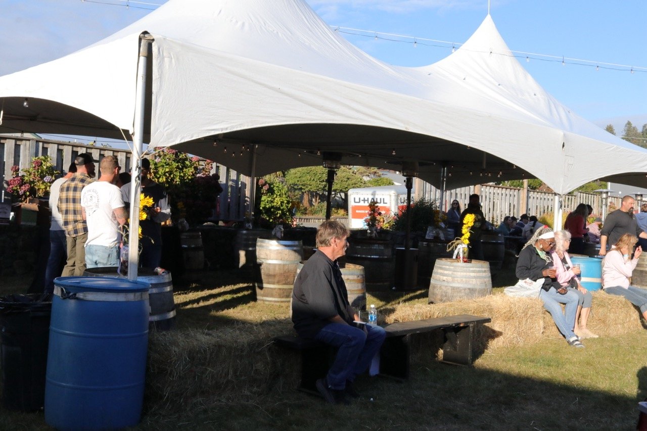 People gathered under a large white canopy at an outdoor event, with some sitting on hay bales and others standing near tables decorated with flowers.