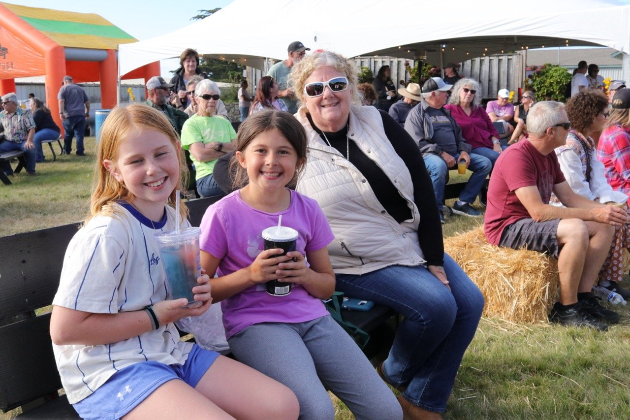 Three smiling females, a young girl with red hair in a baseball jersey, another girl with dark hair in a purple shirt, and an older woman with curly blonde hair and sunglasses, sitting on a bench at an outdoor event during daytime, surrounded by othe