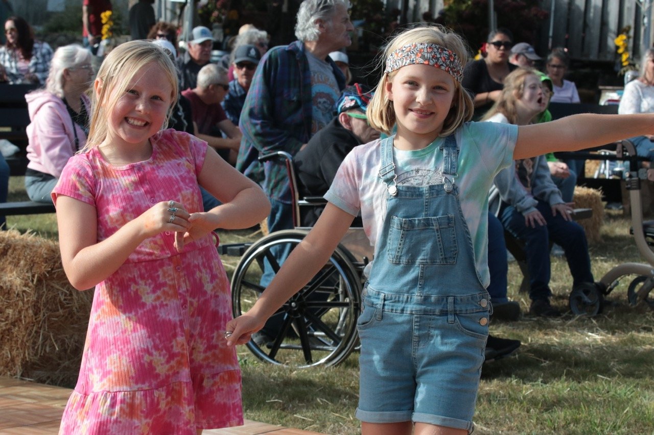 Two young girls smiling and holding hands outdoors at a gathering or festival, with a crowd of seated adults behind them.