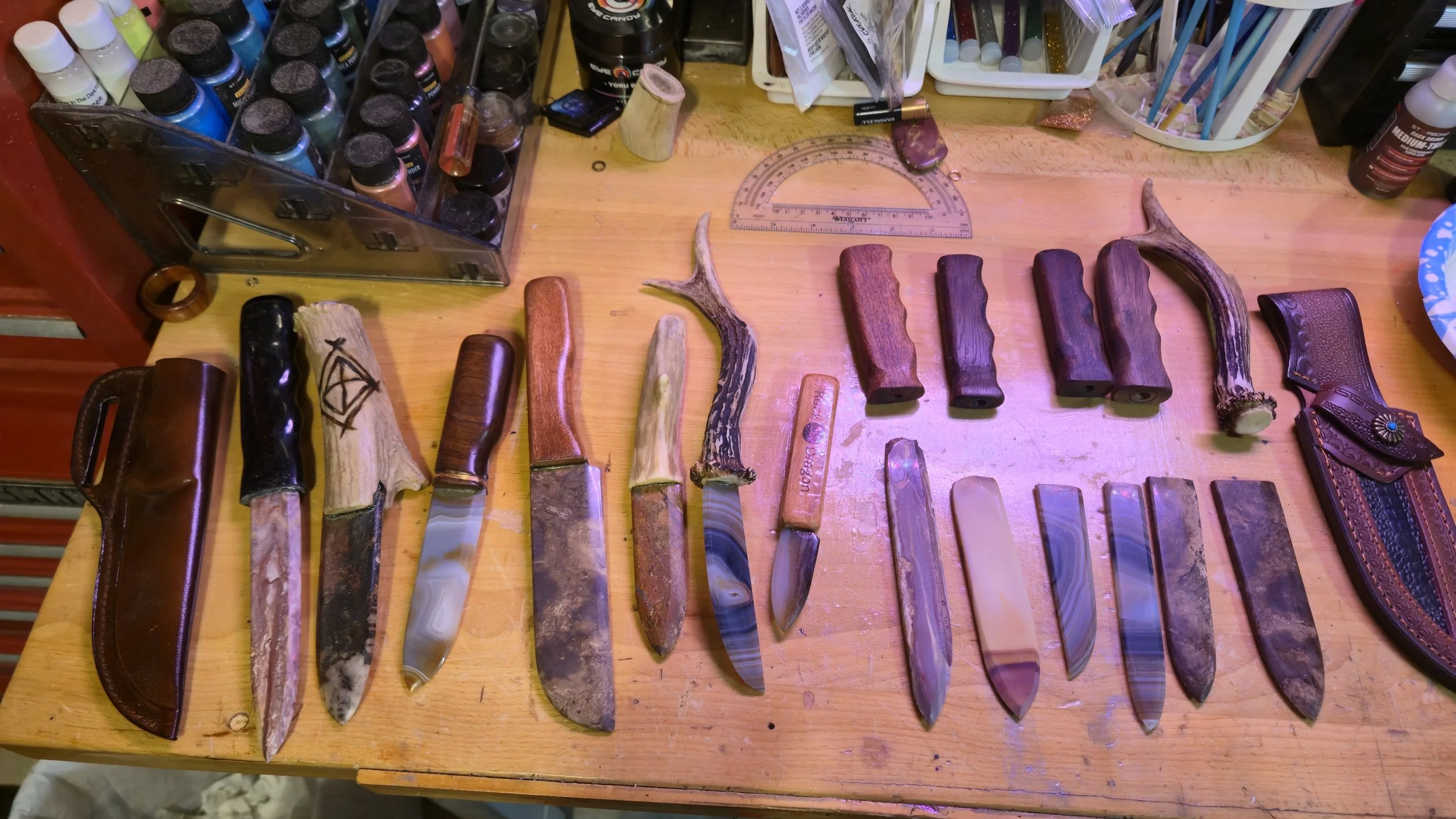 A collection of knives with various handles and blades laid out on a wooden worktable, with leather sheaths, paint bottles, and crafting tools nearby.
