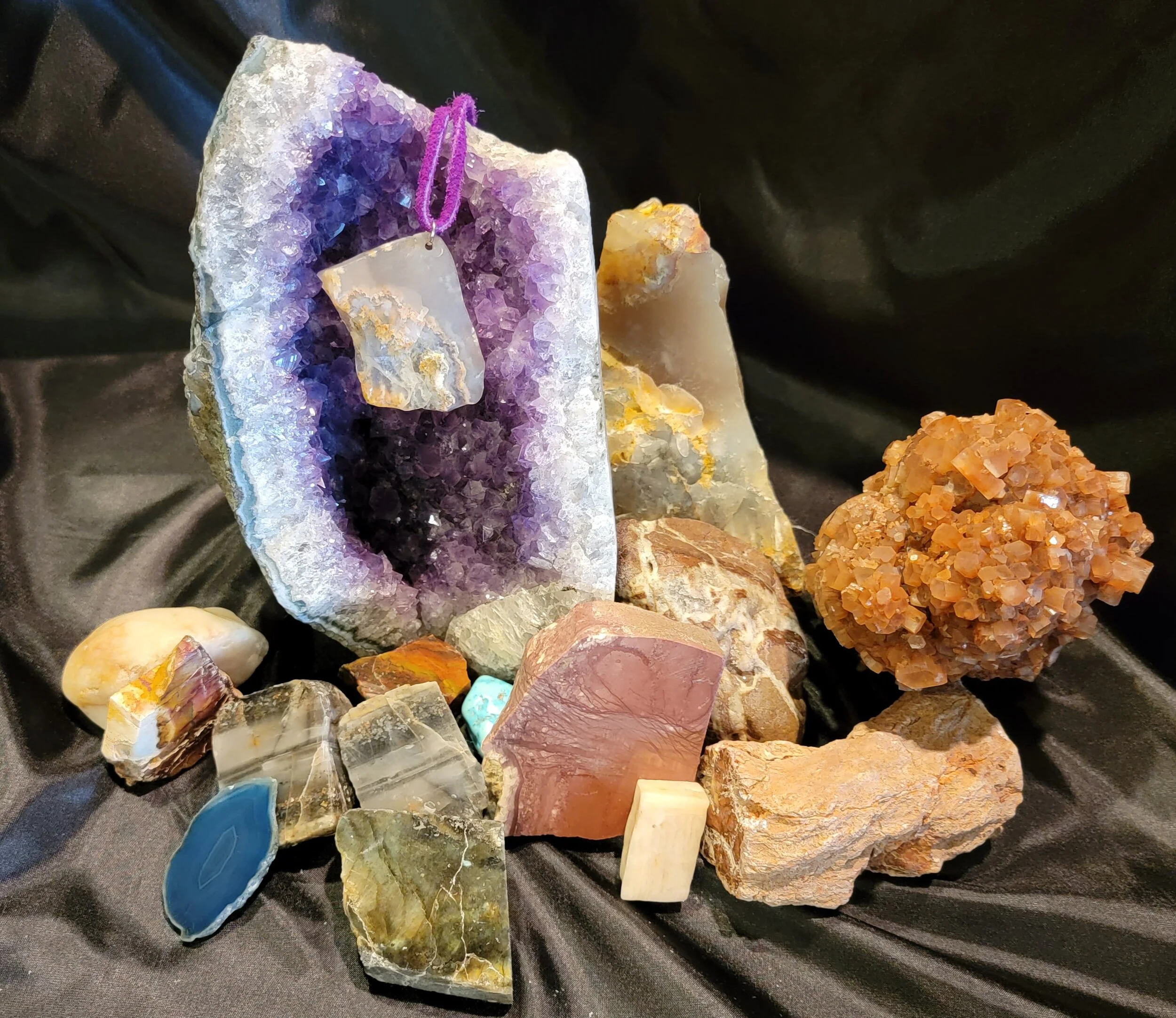 A collection of various rocks and minerals arranged on black fabric, with a large amethyst geode at the center, some with colorful crystals and others with smooth surfaces.