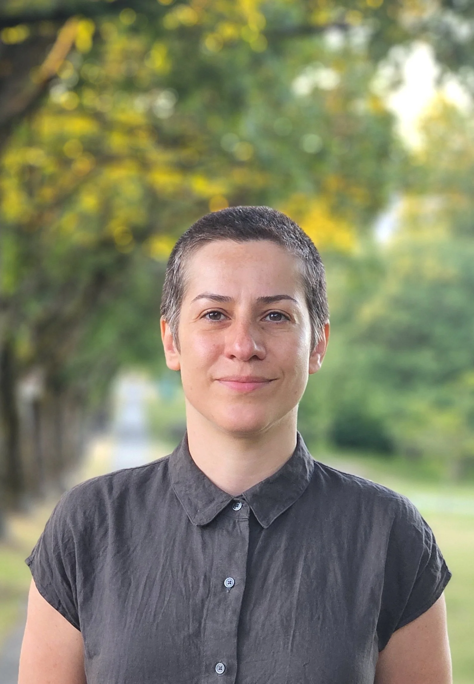 A woman with short dark hair wearing a dark gray button-up shirt standing outdoors with a blurred background of trees and greenery.