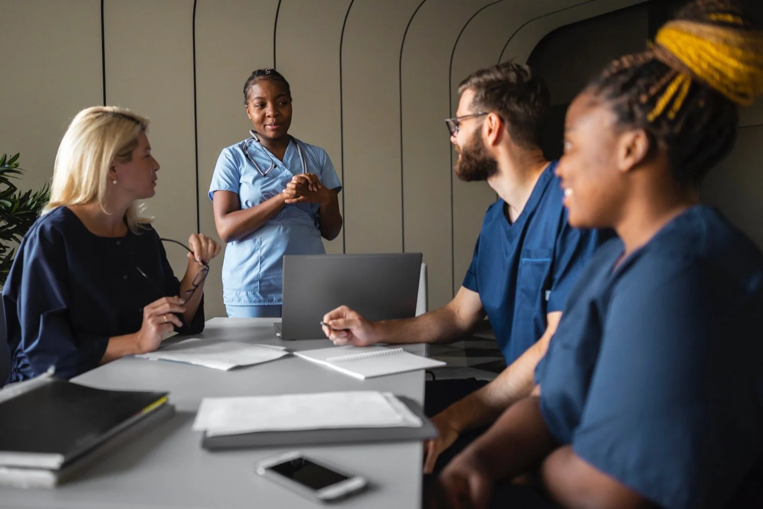 A group of four healthcare professionals sitting around a table in a meeting room, with a nurse standing and speaking to them.