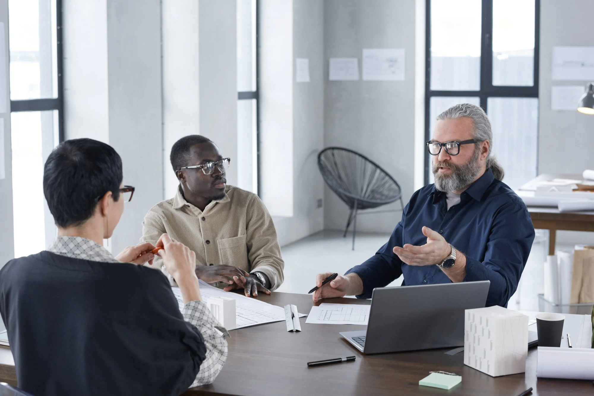Three people having a discussion at a table in a modern office with large windows.