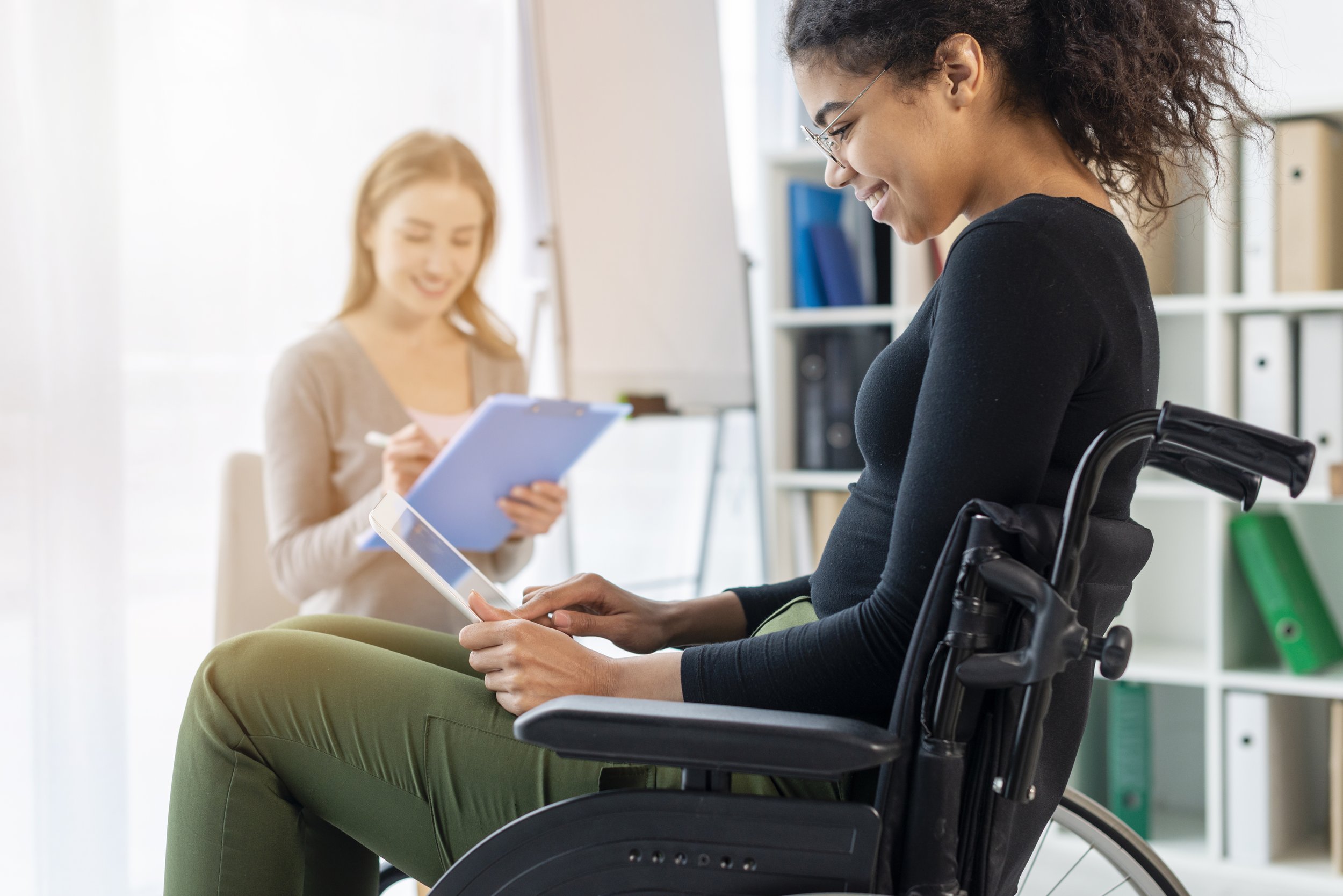 A woman in a wheelchair using a tablet while another woman notes on a clipboard in an office setting.