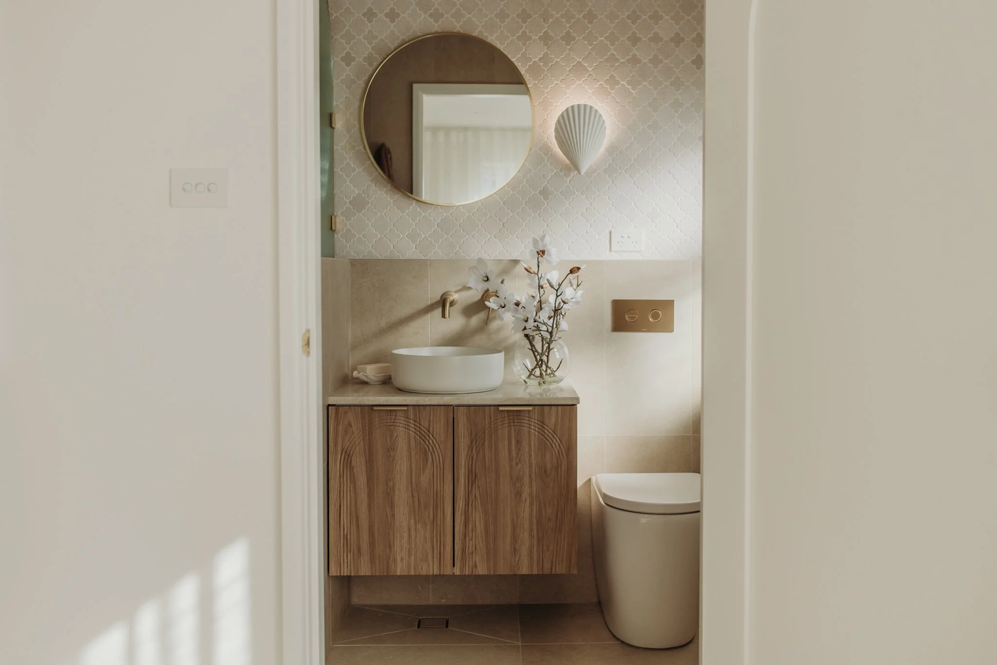 A modern bathroom with a wooden vanity, white vessel sink, round mirror, wall-mounted faucet, vase with white flowers, wall-mounted toilet, and decorative wall light.