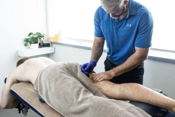 An osteo wearing a blue shirt and gloves performing dry needling on a person lying face down on a massage table covered with a towel. There's a small table with plants in the background.