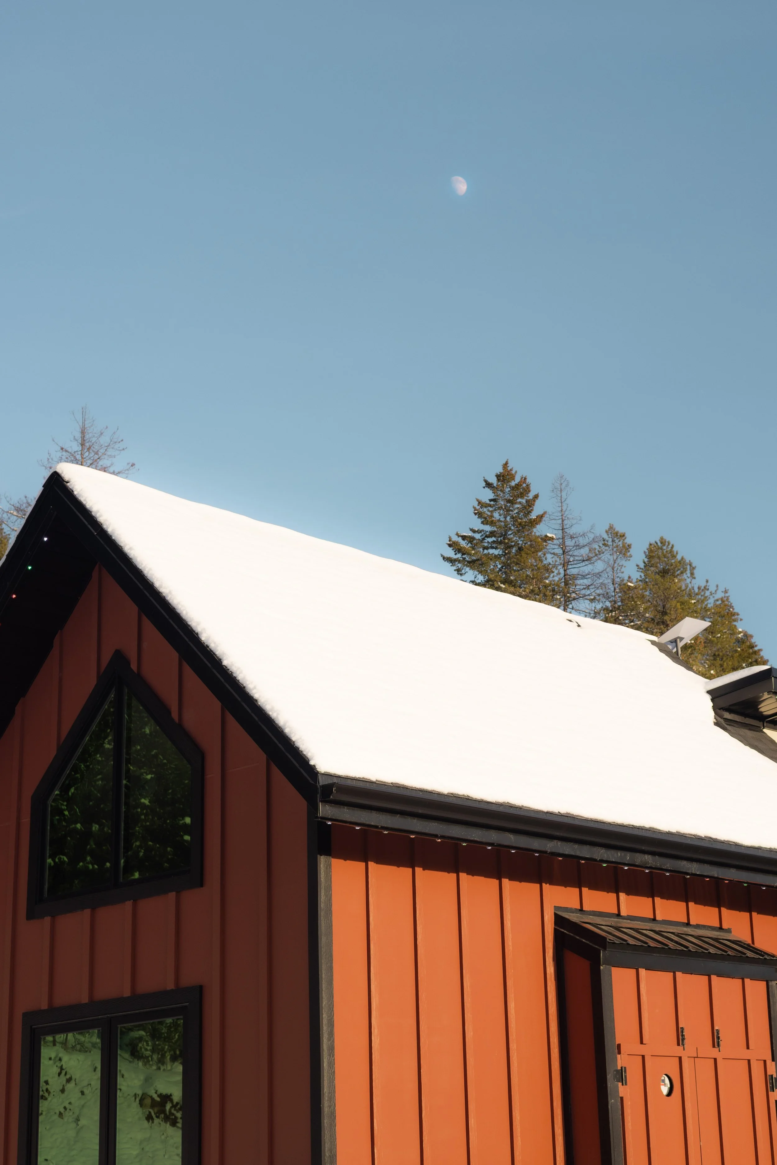 A house with an orange exterior and black window frames, snowy roof, under a clear blue sky with visible moon and trees in the background.