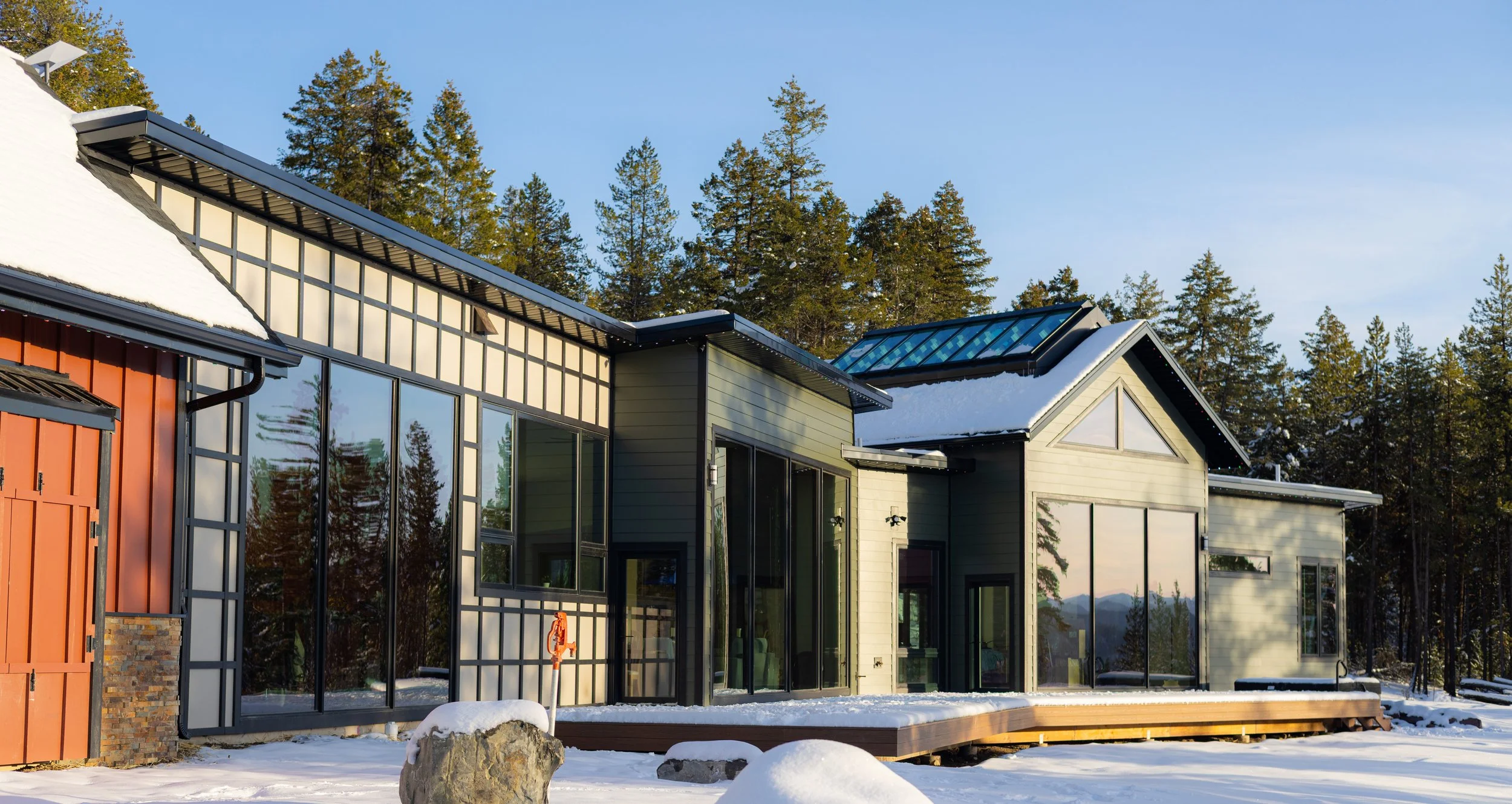 Modern house with large glass windows surrounded by snow, trees, and a clear sky.