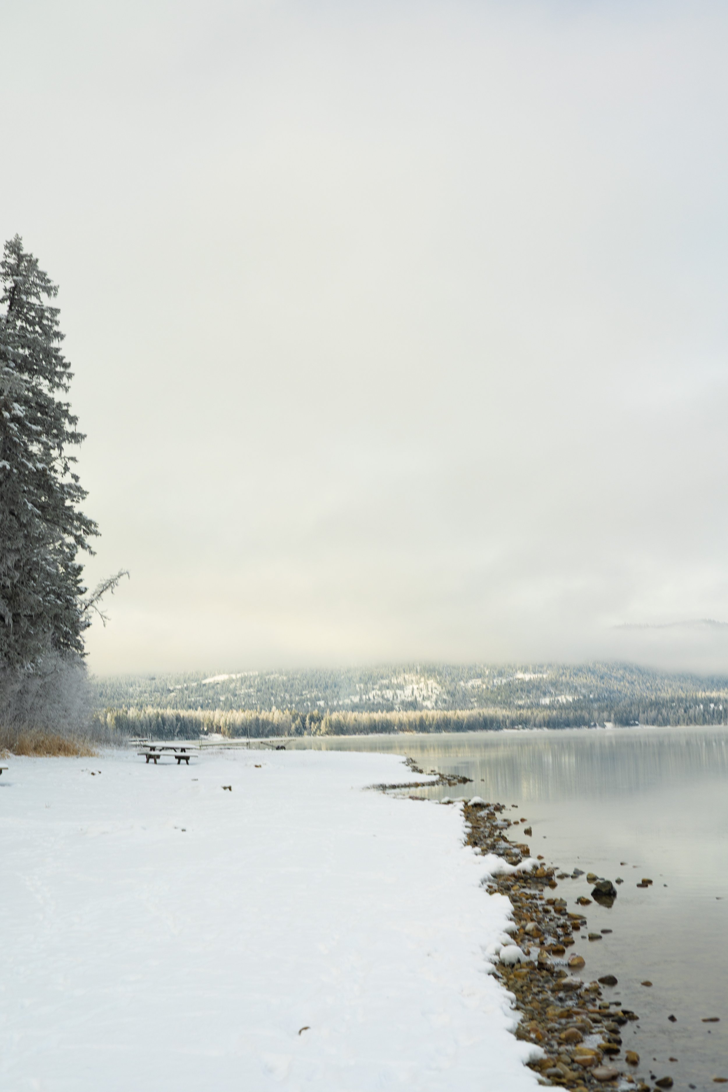 Snow-covered shoreline beside a calm body of water with a forested hill in the distance and a cloudy sky overhead.