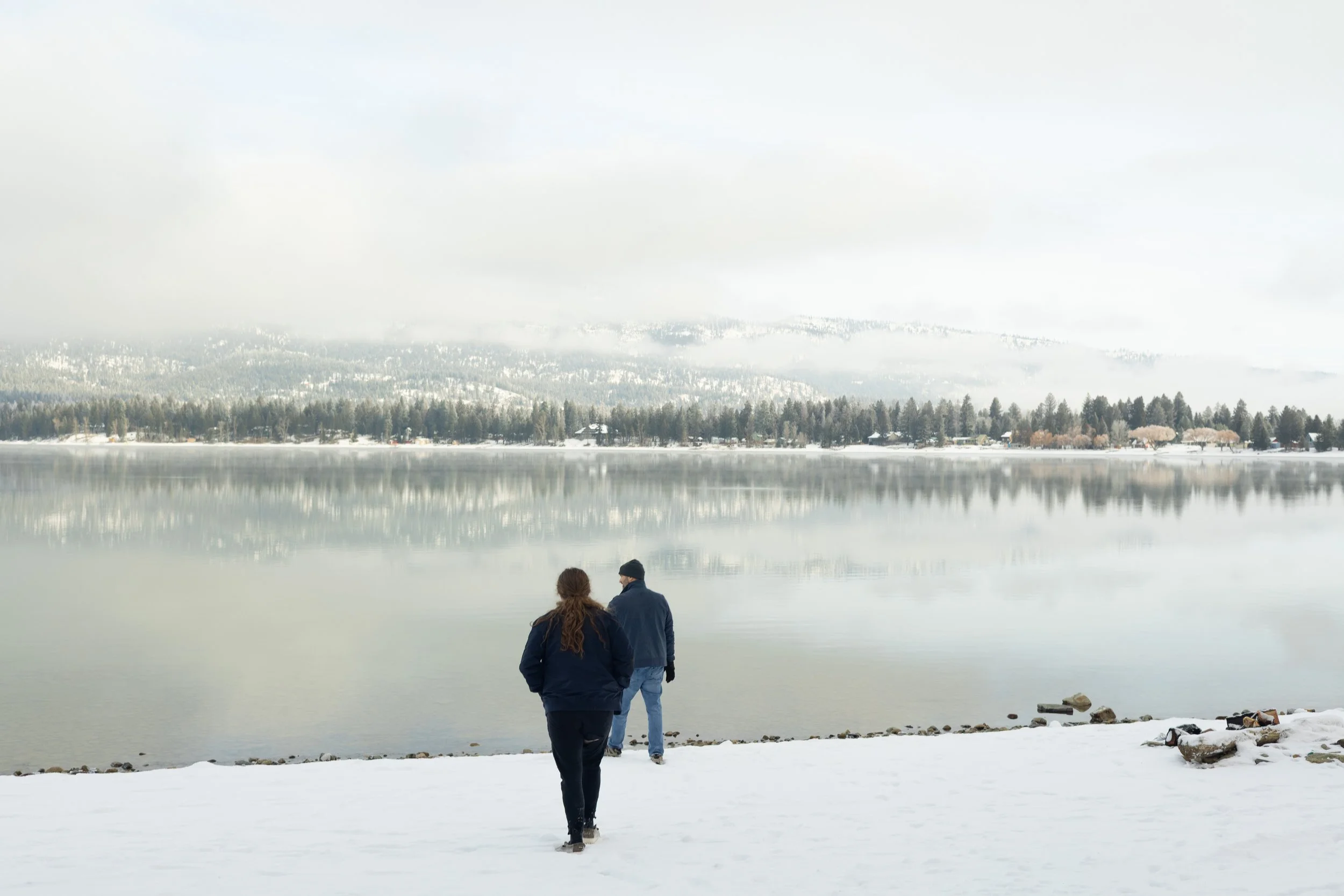 Two people walking along a snowy lakeshore, with a calm lake and a snow-covered landscape in the background.
