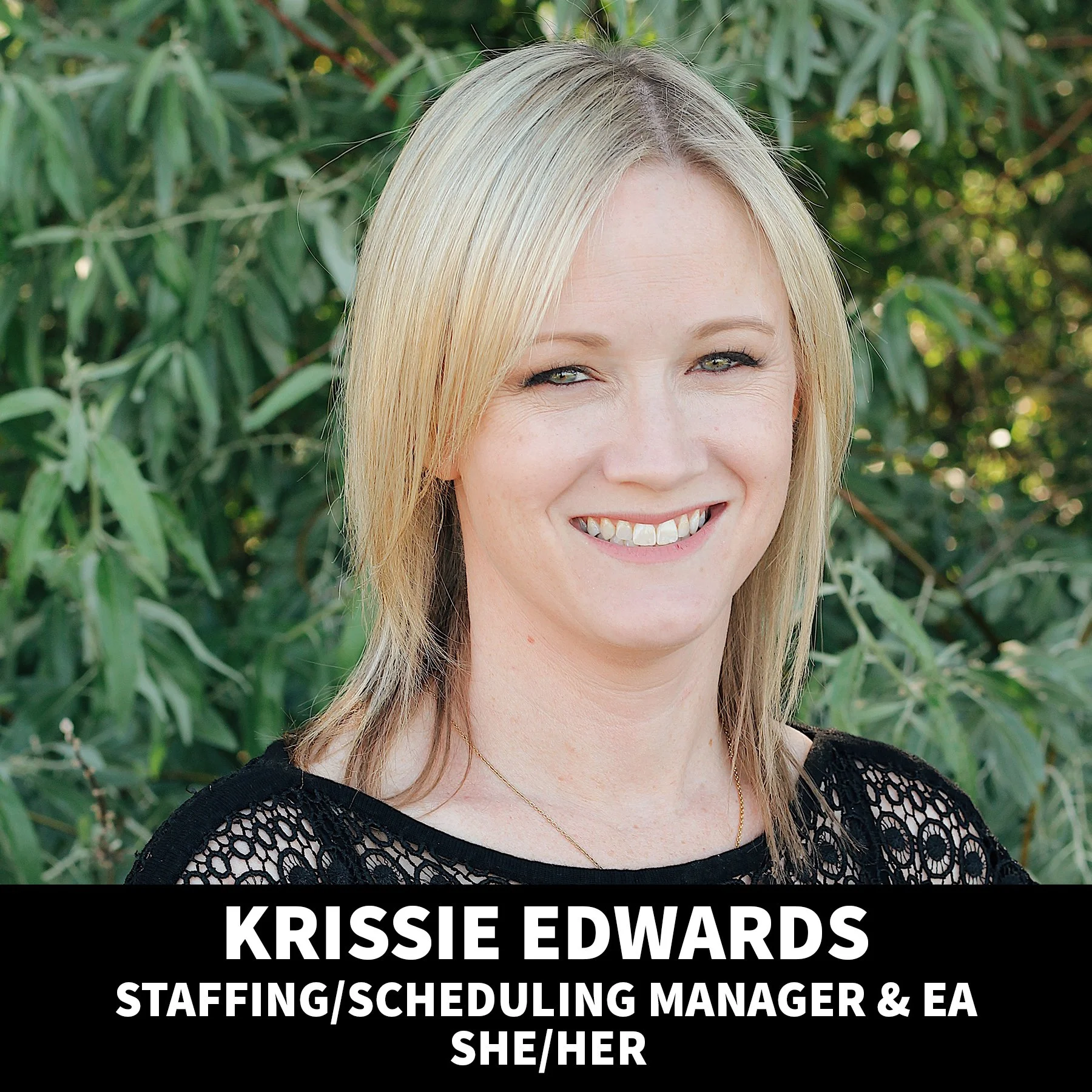 A smiling blonde woman with light makeup, wearing a black top, standing outdoors with green foliage in the background.