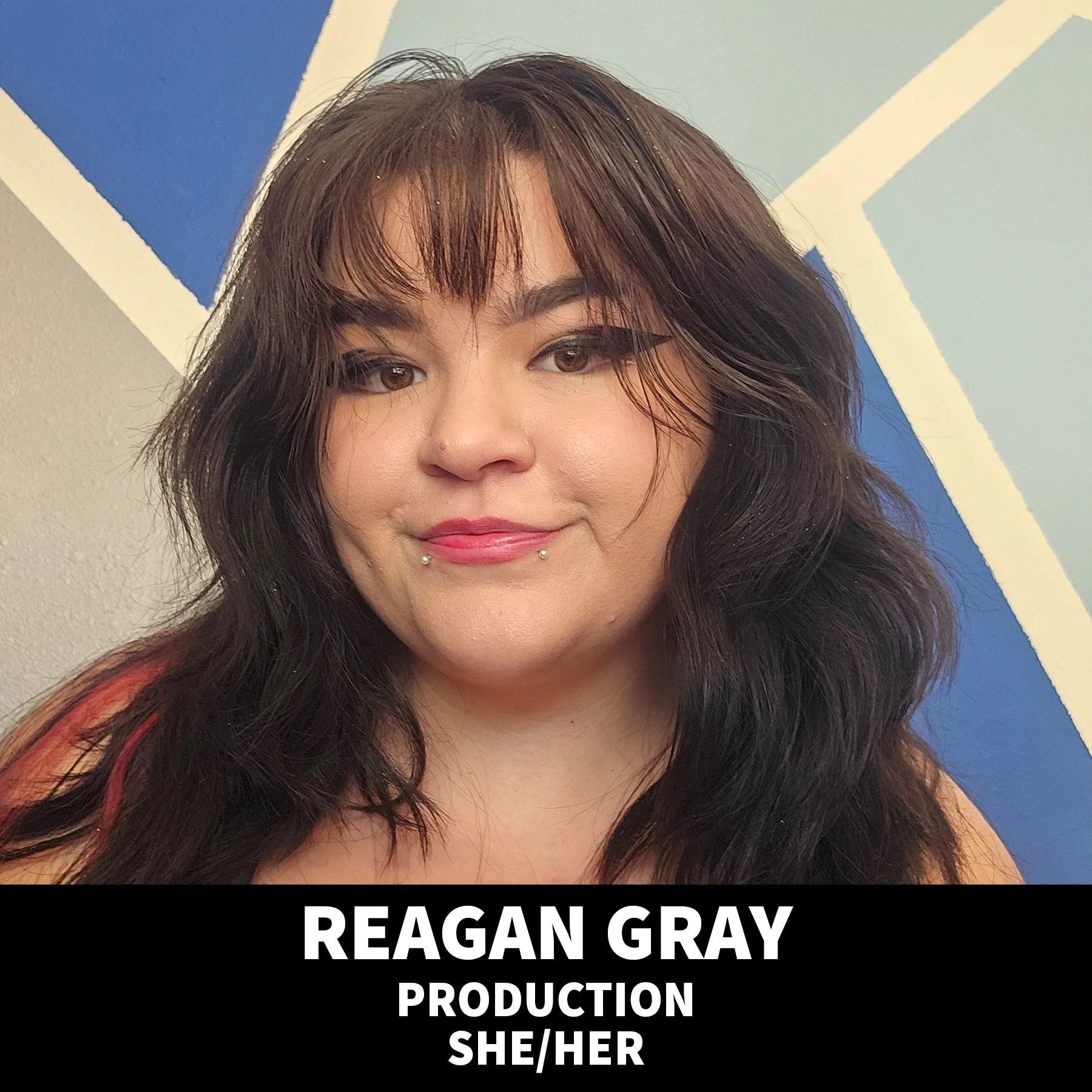 A woman with long dark hair smiling and wearing makeup in front of a beige background, with a black banner at the bottom that says 'Reagan Gray, Production, She/Her.'