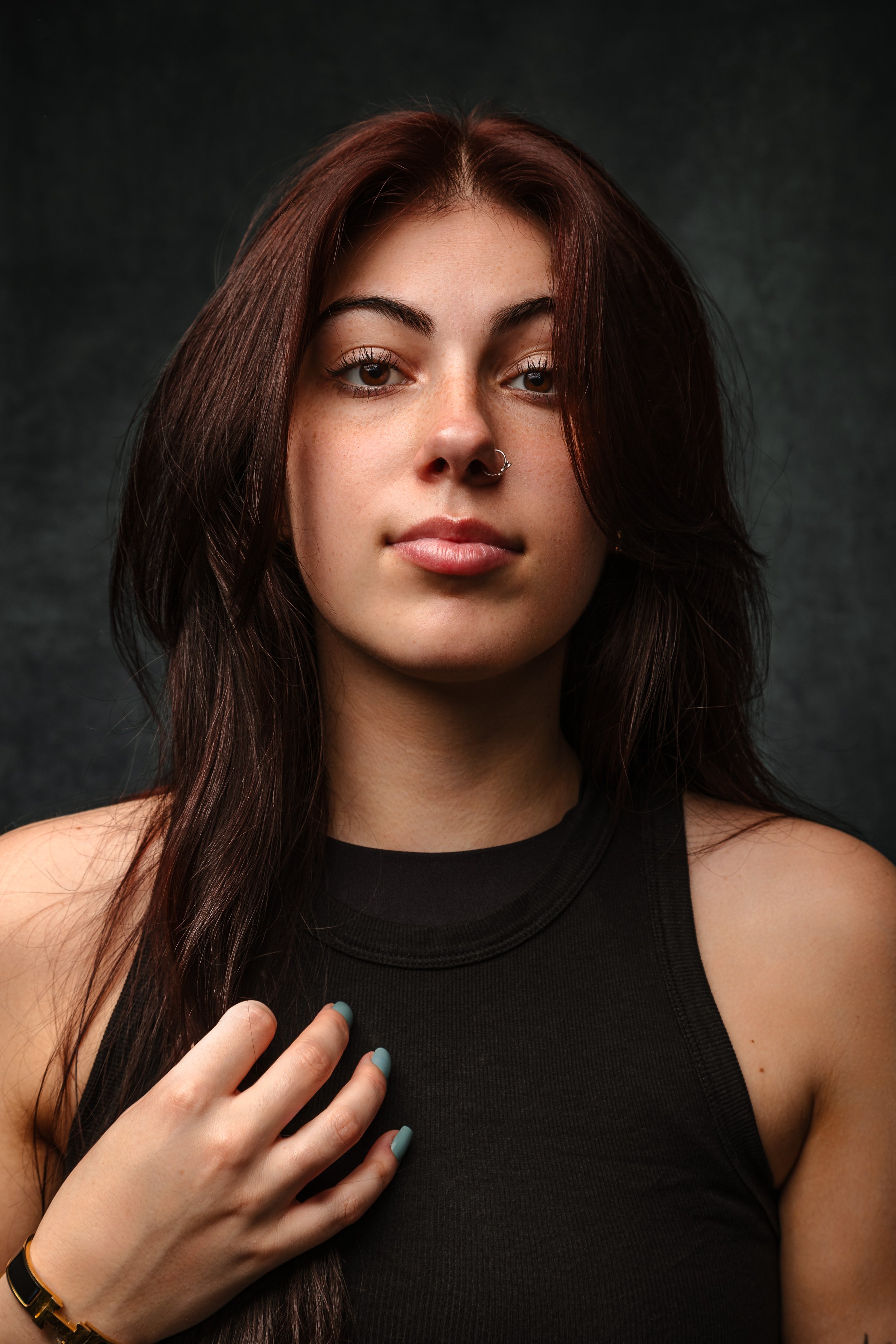 Woman with long dark hair, wearing a black top, and has a nose piercing; hand on chest with painted nails, dark background.