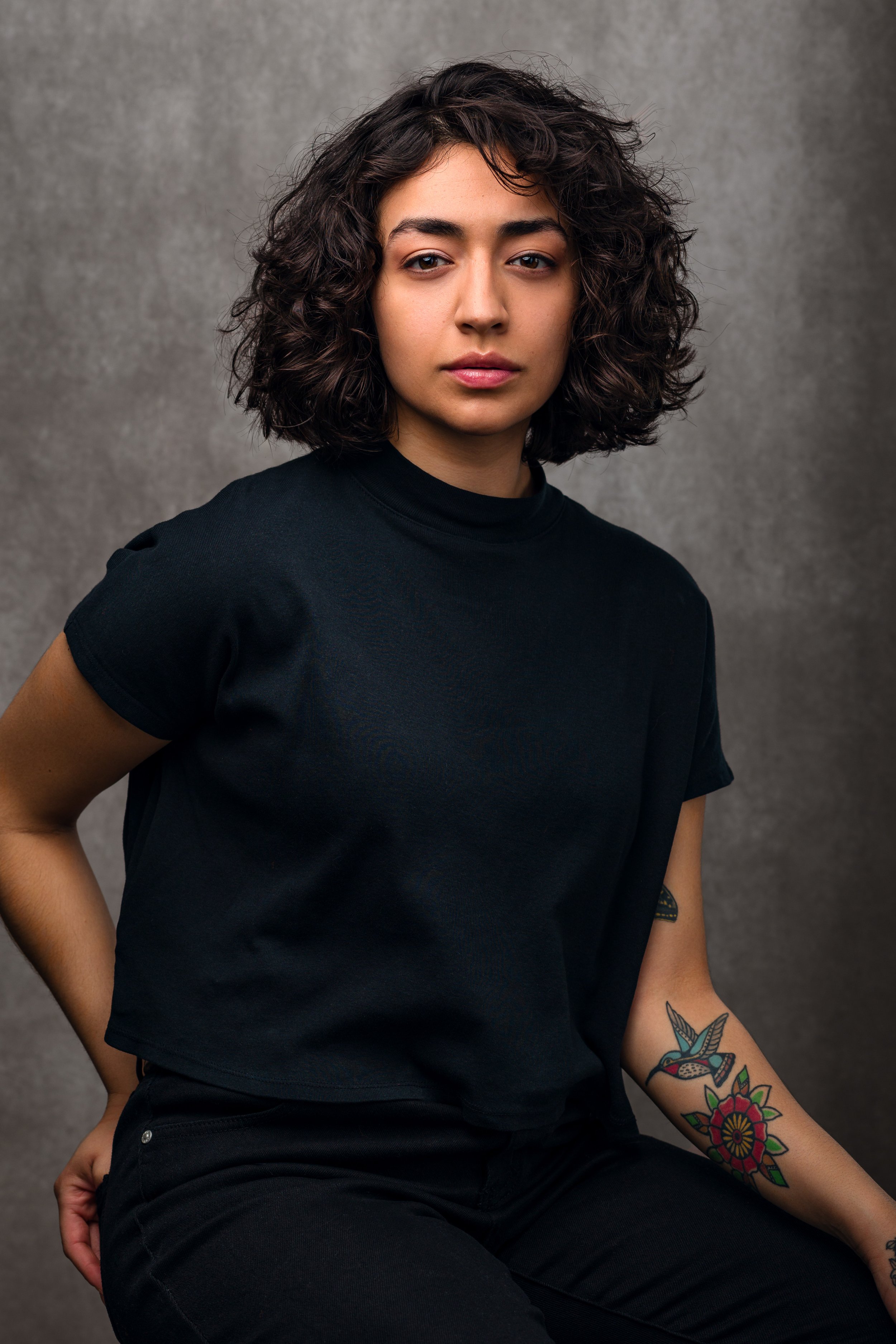 Person with curly hair and tattoo, wearing a black shirt, seated against a gray background.