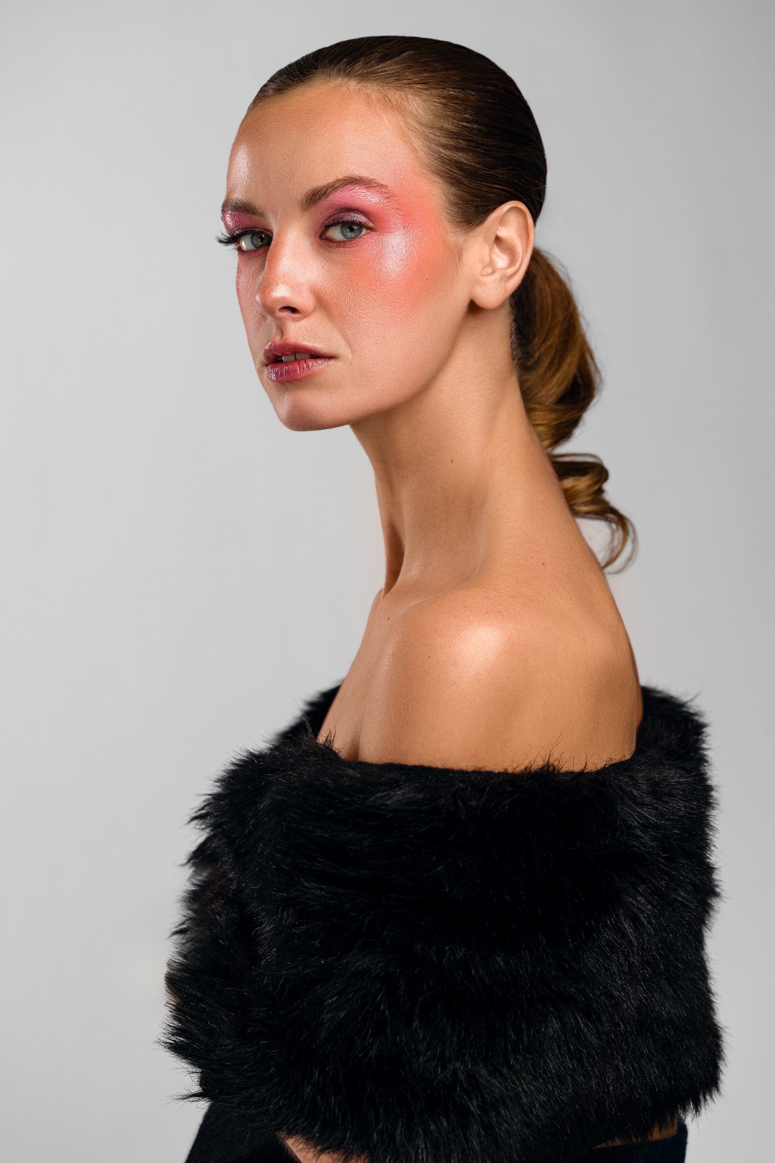 Profile portrait of a woman wearing dramatic pink makeup, with her hair tied back, dressed in a black fur wrap, against a plain background.