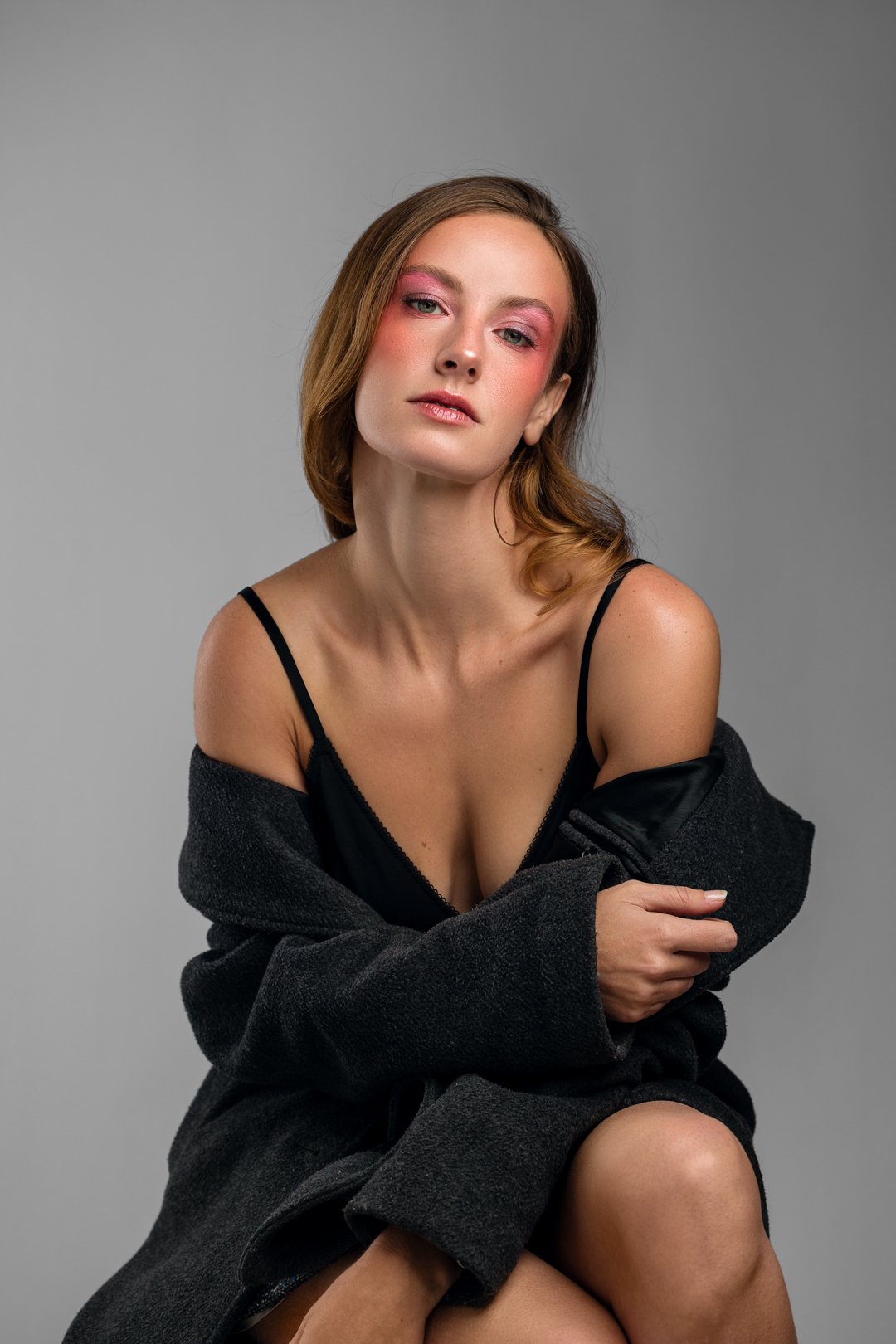 Woman with pink makeup wearing a black dress and gray coat seated against a gray background