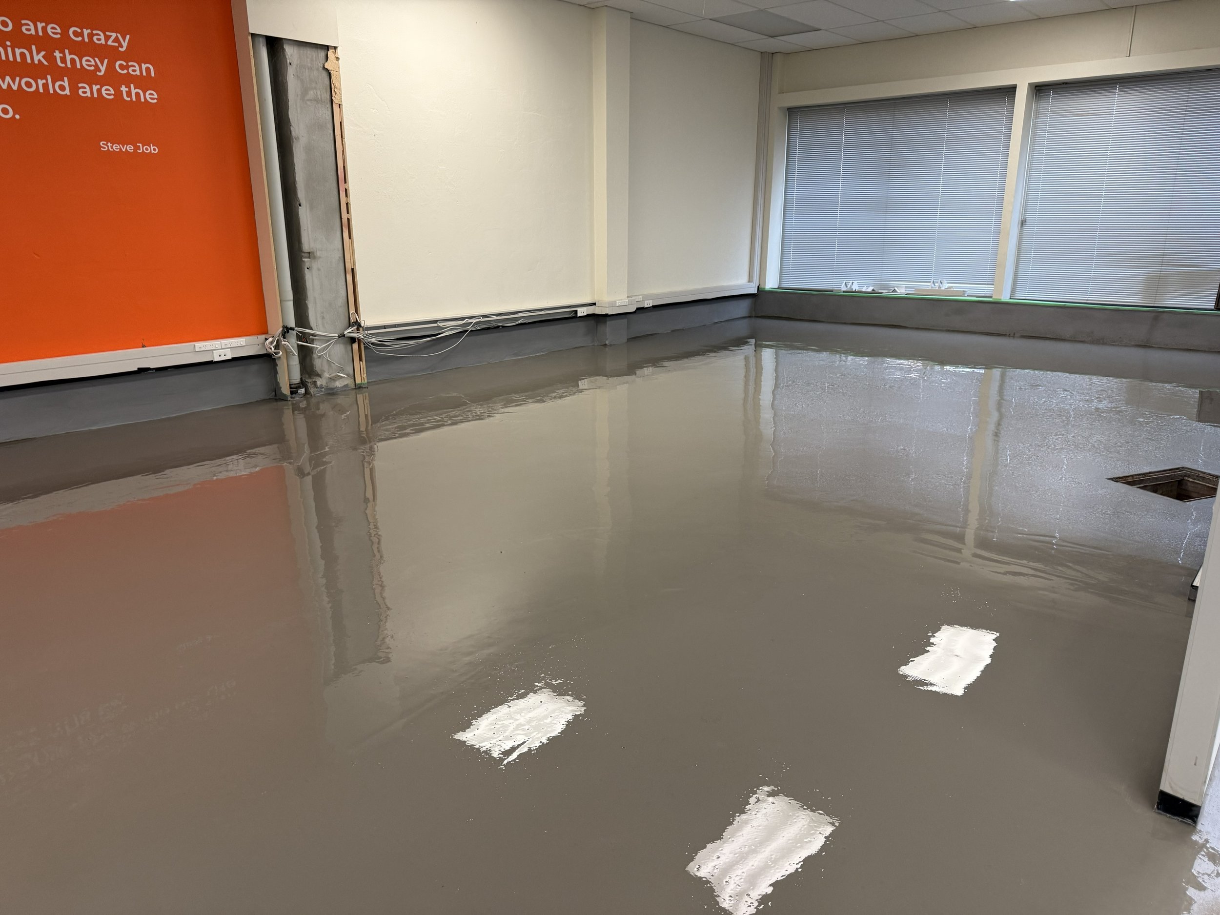 An empty office space with large windows a concrete floor with self leveling compound still wet from recent flooring work, and a partially removed wall with visible wiring and insulation.