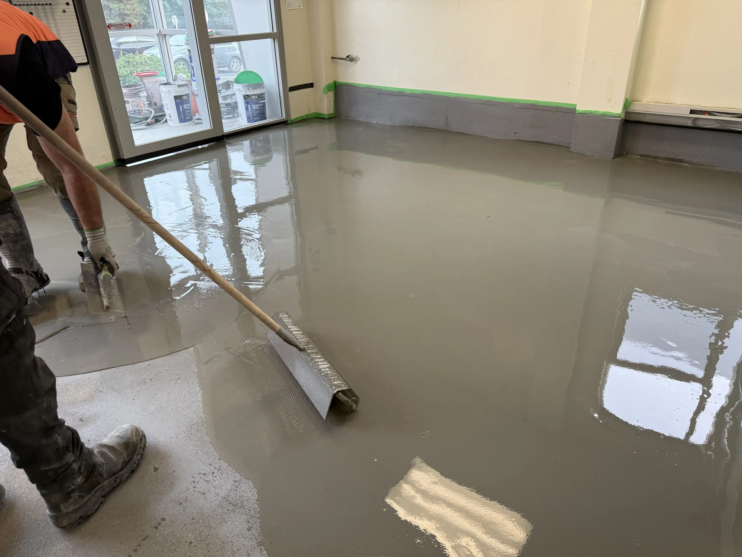 Worker smoothing freshly poured self leveling compound inside a building near large windows and glass door.