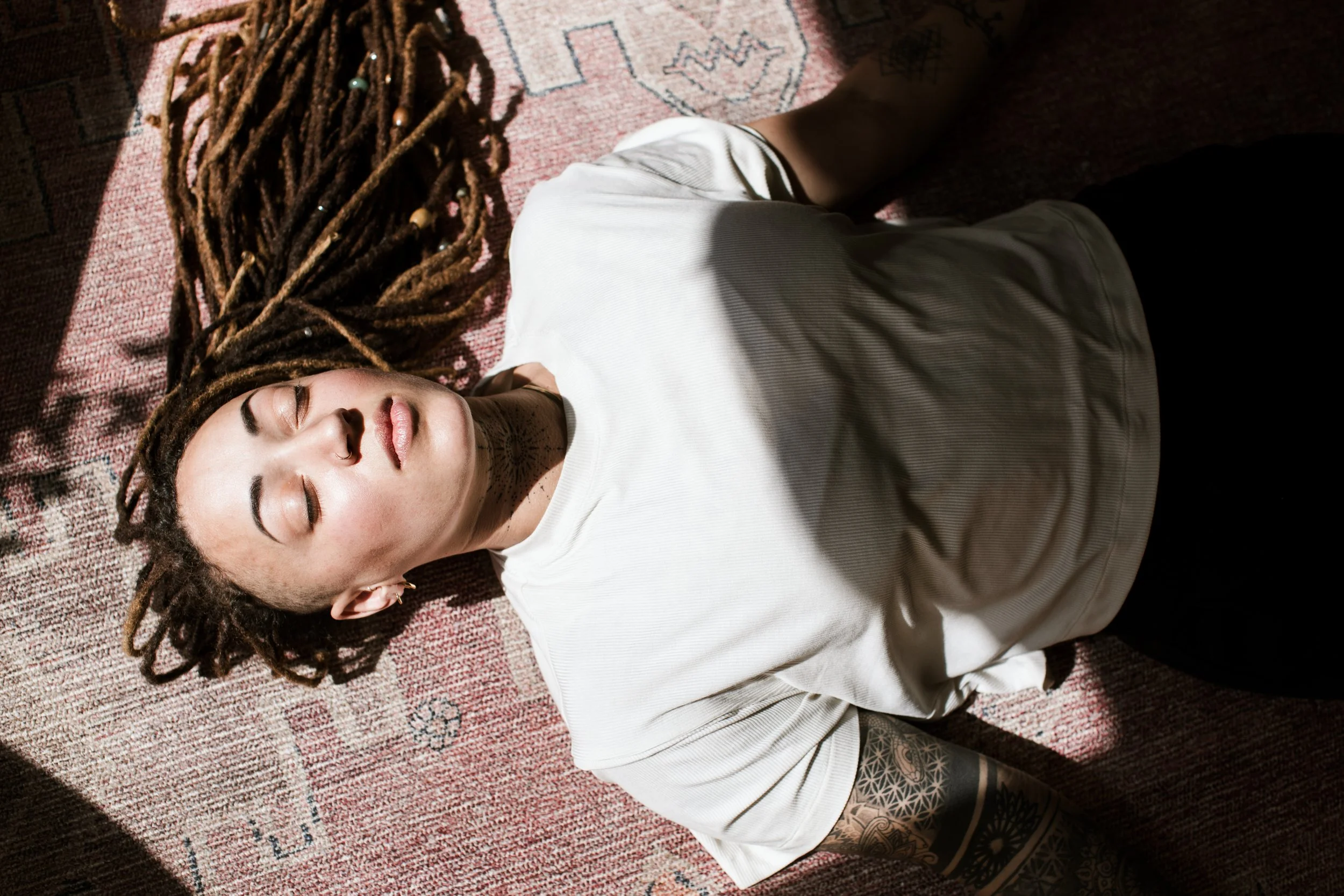 A woman with dreadlocks and tattoos lying on a patterned rug with her eyes closed, wearing a white t-shirt.