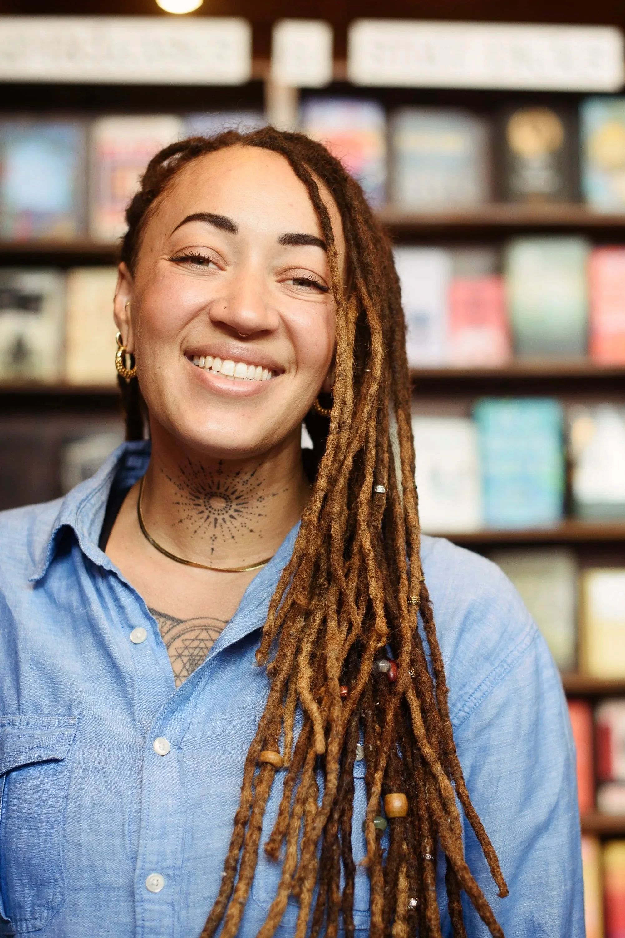 Smiling woman with dreadlocks and tattoos in front of a bookshelf.
