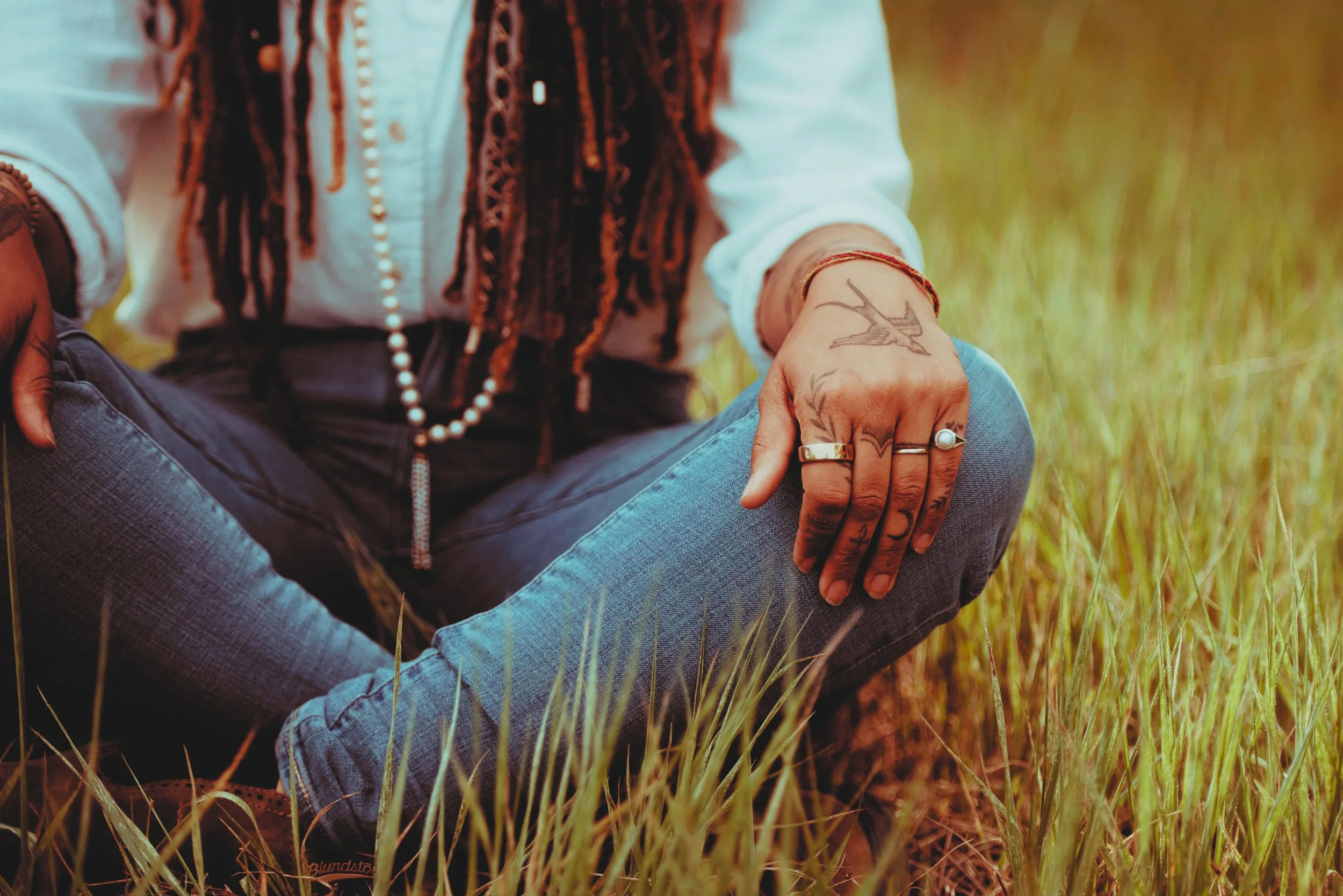 A meditation teacher sitting cross-legged in a grassy field, wearing a white shirt and blue jeans, with tattoos and rings on their fingers, and a beaded necklace around their neck.