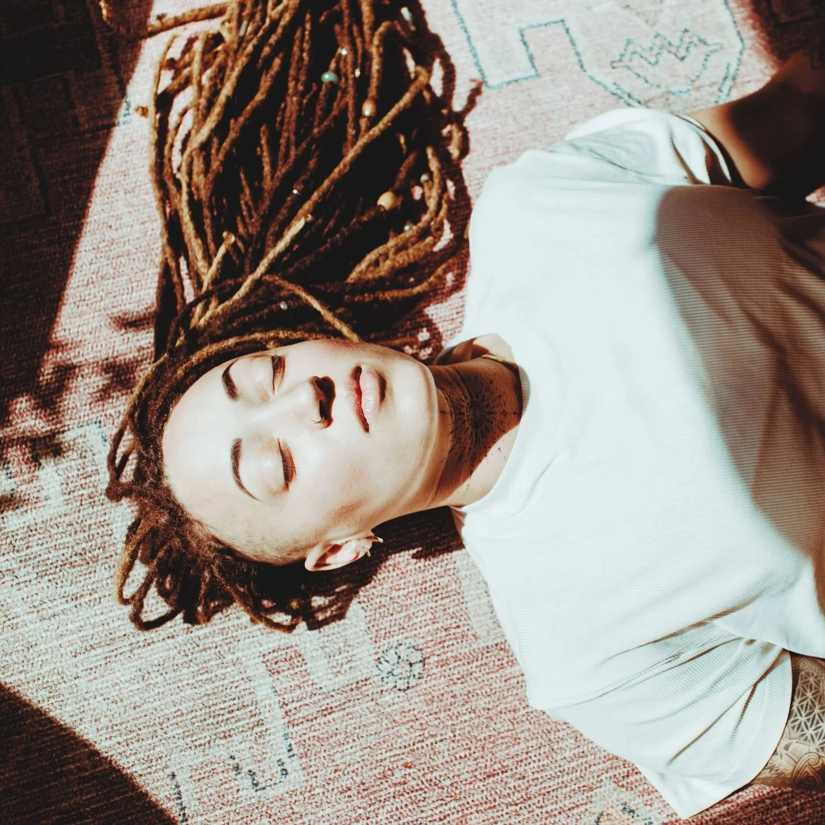 A meditation teacher with dreadlocks lying on a patterned rug with closed eyes, wearing a white shirt, and a tattoo on their neck visible, teaching the an advanced meditation technique