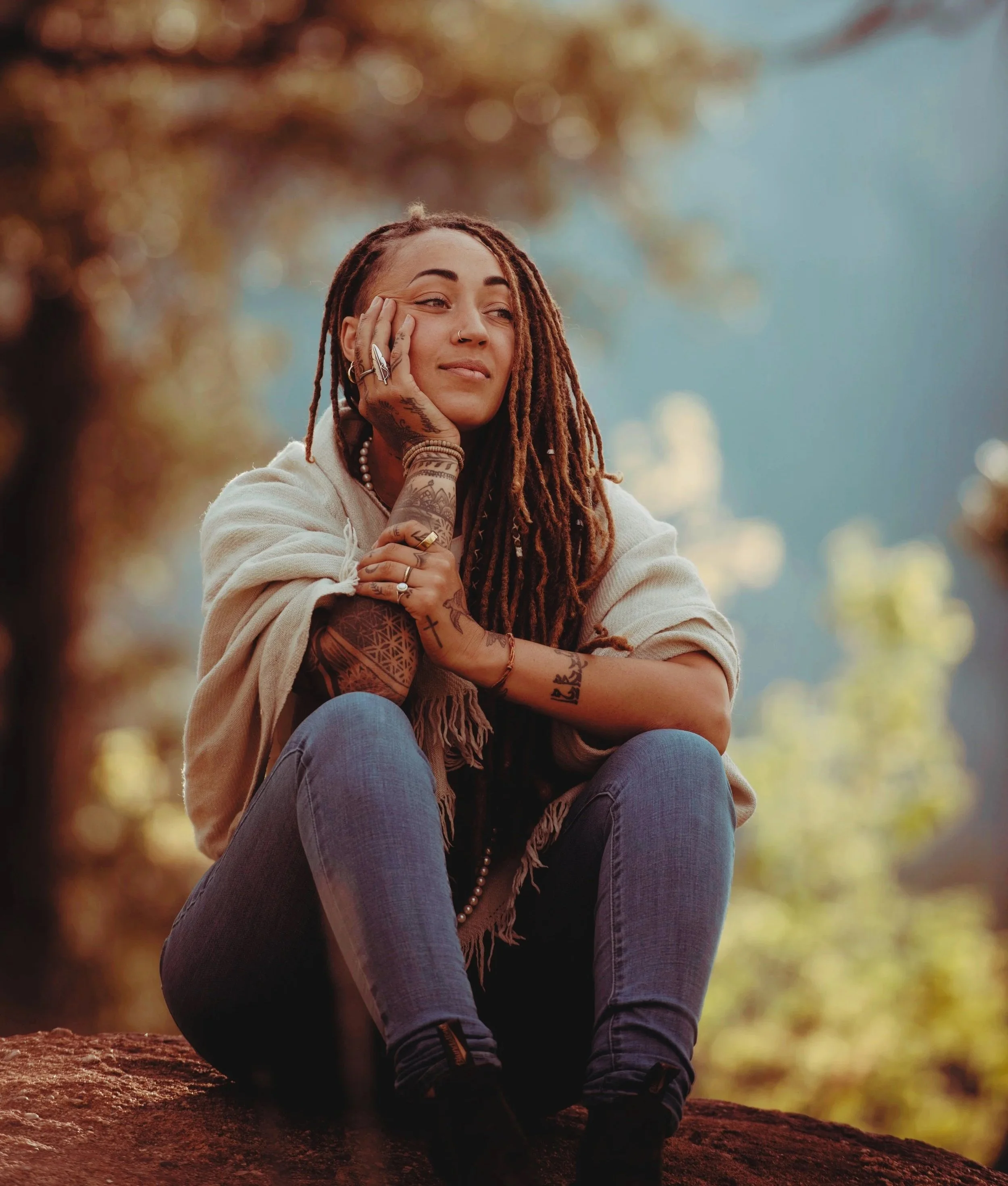 A meditation teacher with dreadlocks sits outdoors on a rock, resting her face on her hand, with trees and a blue sky in the background.