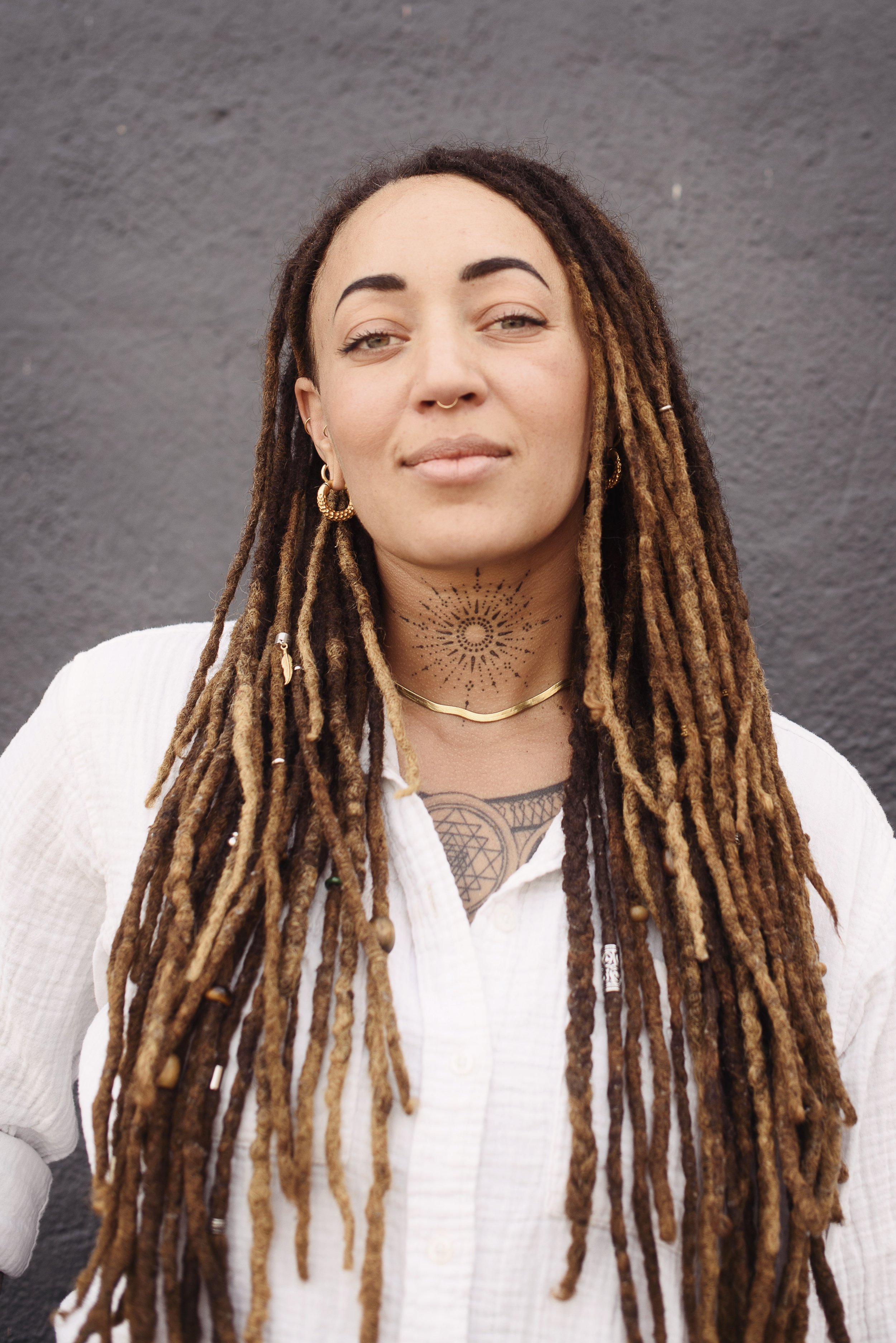 A woman with long, brown dreadlocks wearing gold earrings, a gold necklace, and a white button-up shirt with a black tattoo on her chest, standing against a dark gray wall.