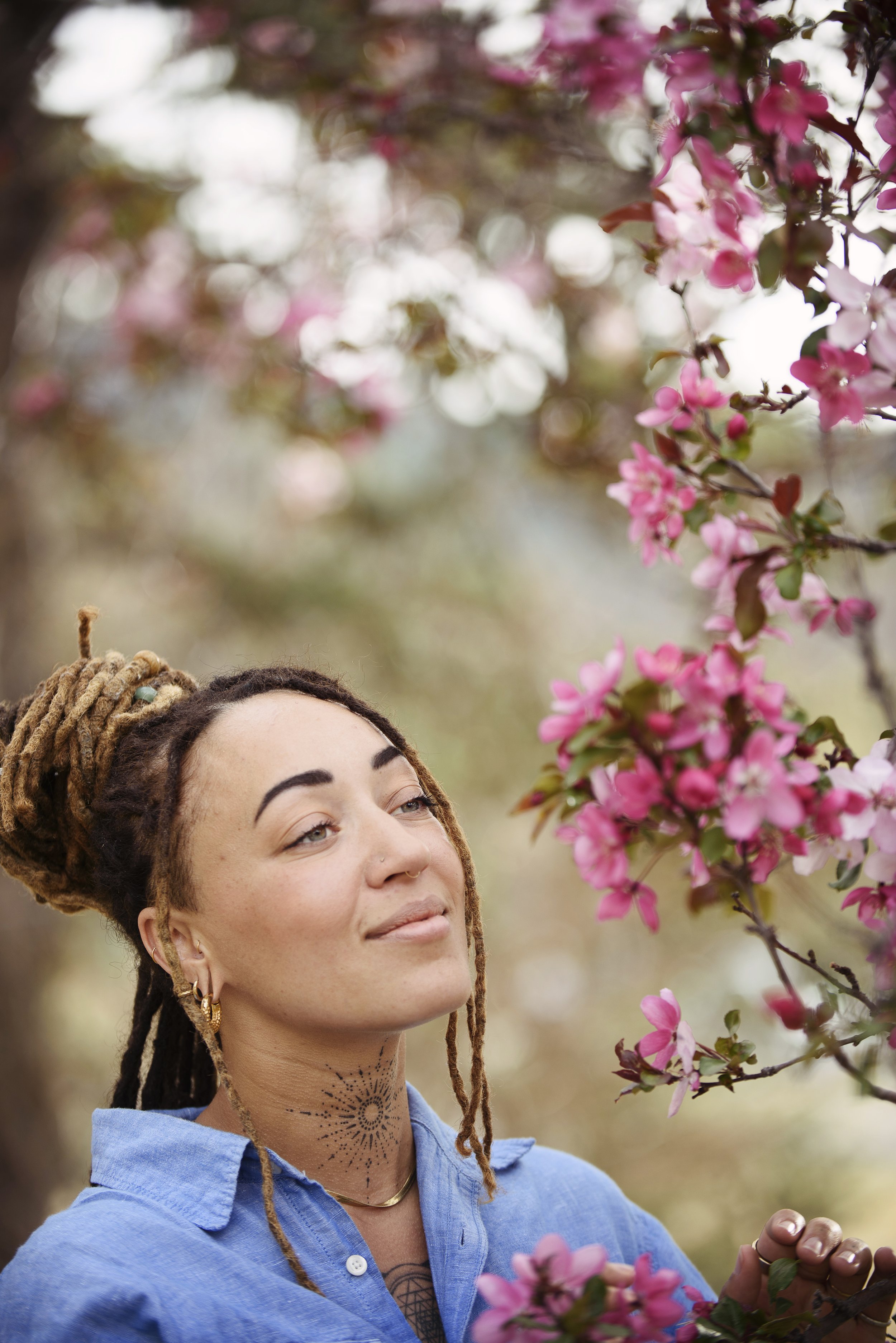 A woman with dreadlocks and tattoos standing among pink blossom trees, looking thoughtful and serene.