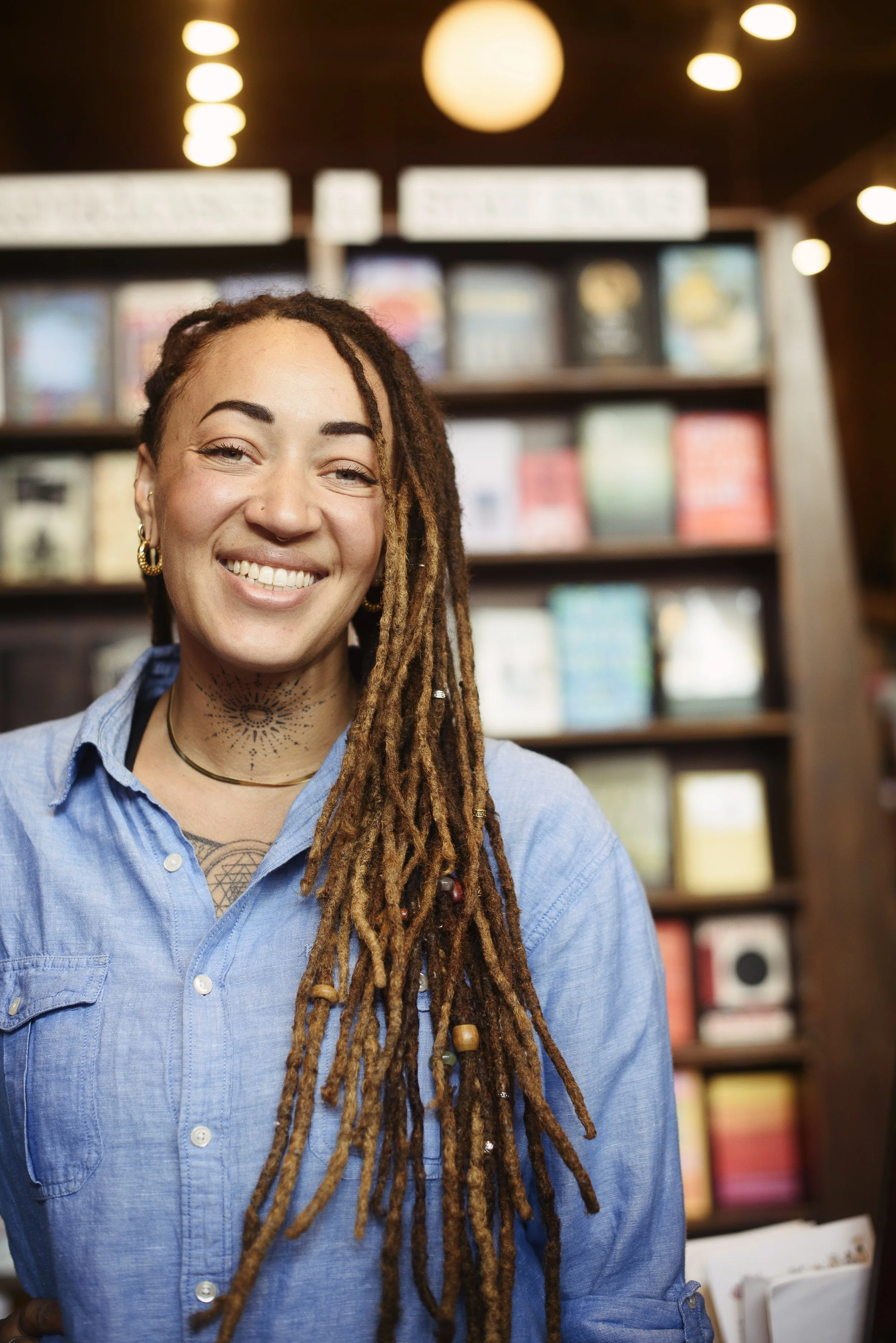 A woman with long dreadlocks, smiling, wearing a blue denim shirt, standing in front of bookshelves in a bookstore.