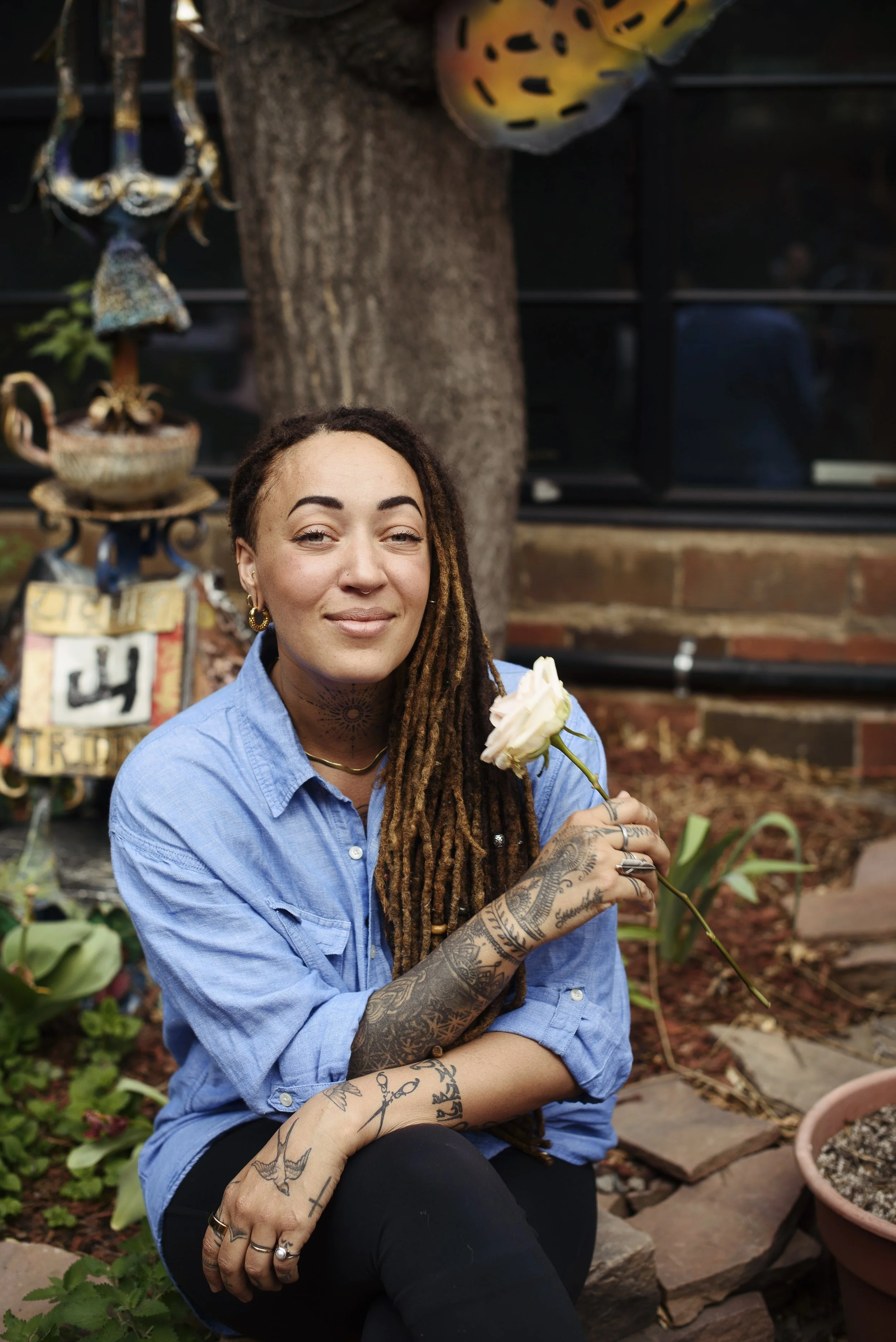 A woman with long dreadlocks, tattoos, and jewelry, wearing a blue shirt, sitting outside holding a white flower, smiling at the camera.