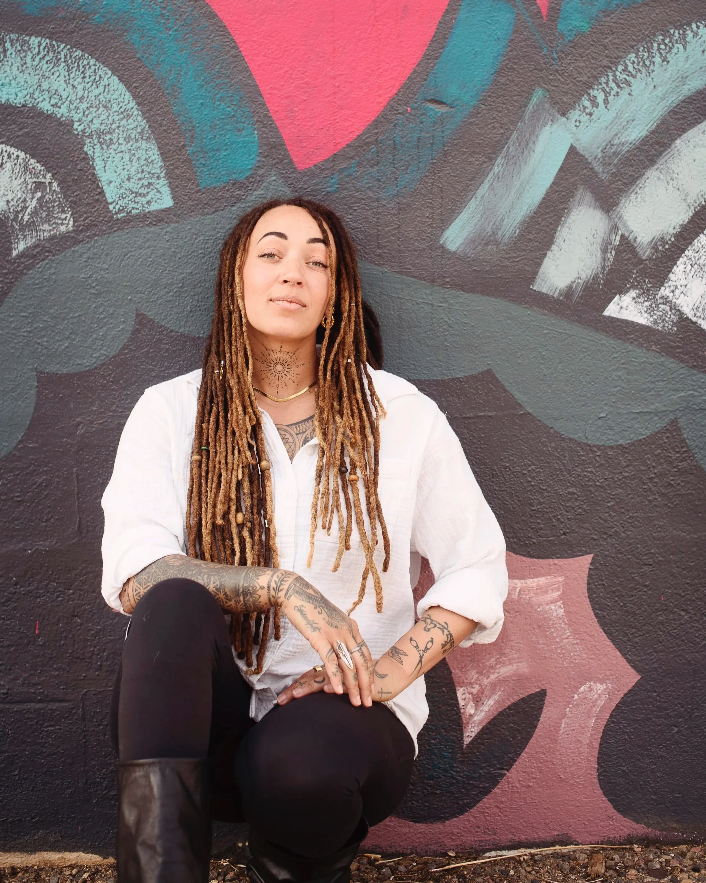 A woman with long dreadlocks, tattoos on her arms and chest, wearing a white shirt and black pants, crouching in front of a colorful graffiti wall.