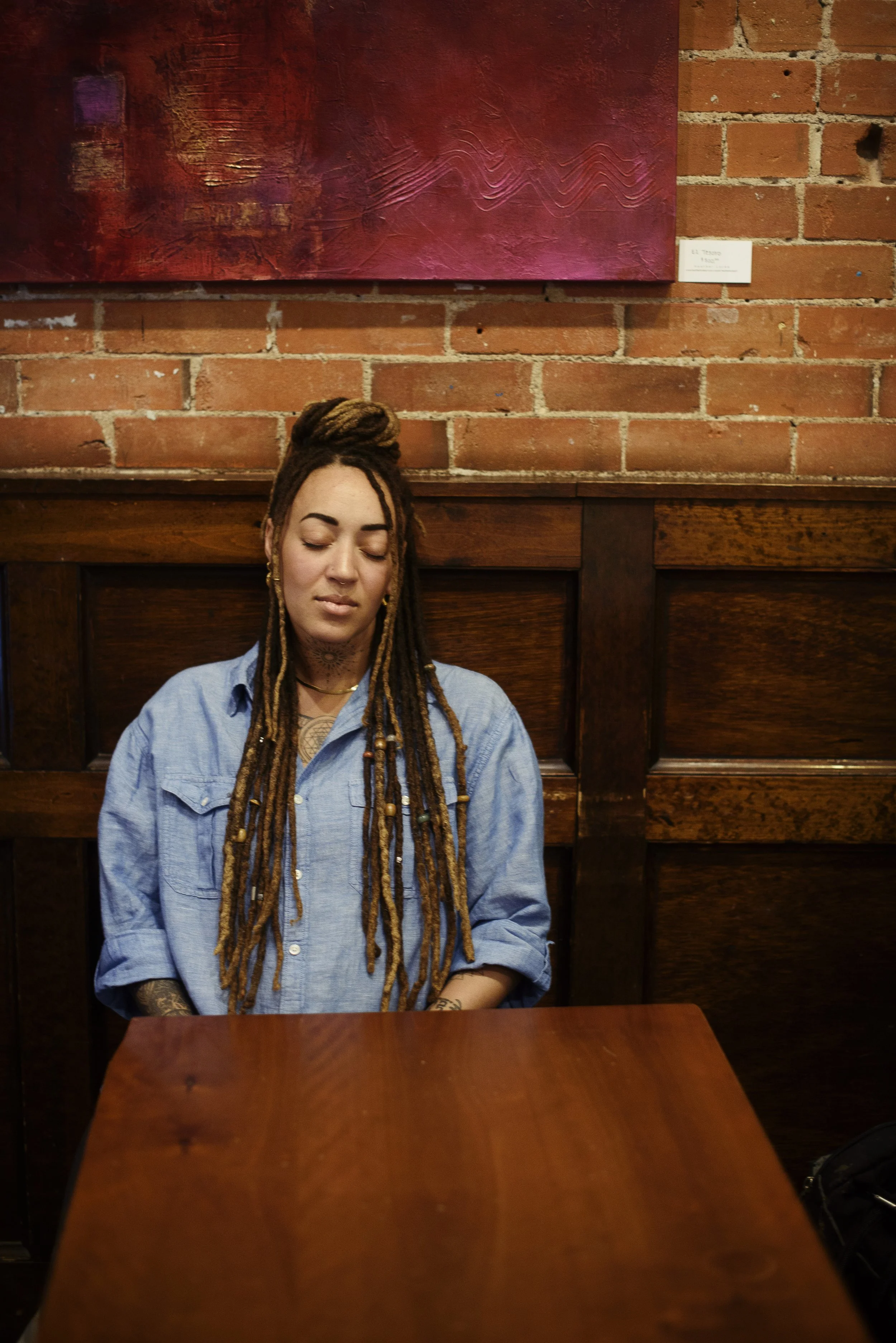 A woman with long dreadlocks and tattoos, wearing a blue denim shirt, sitting with closed eyes at a wooden table inside a brick-walled room with a large red abstract painting on the wall.