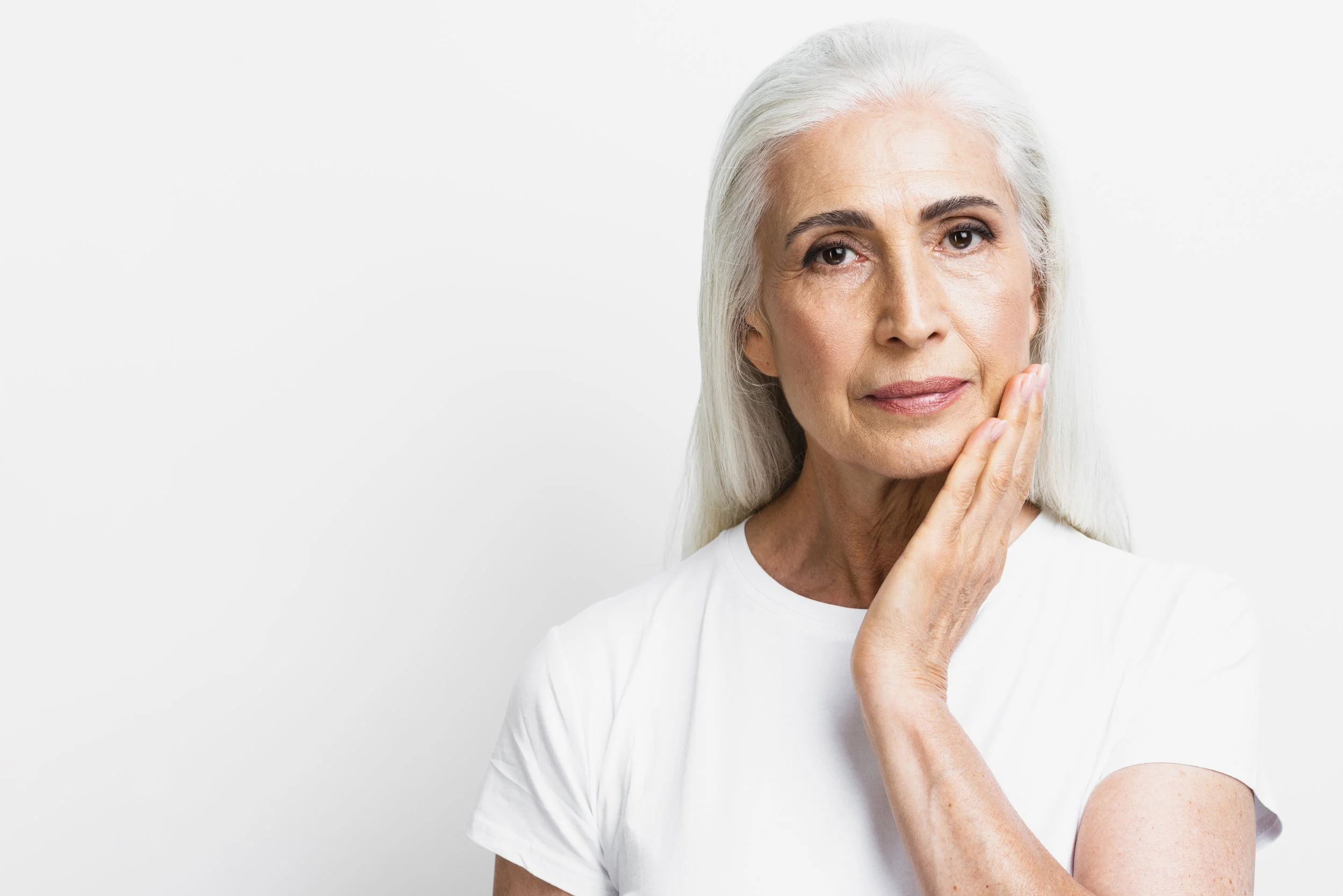 Older woman with long gray hair touching her face, wearing a white shirt, on a white background.
