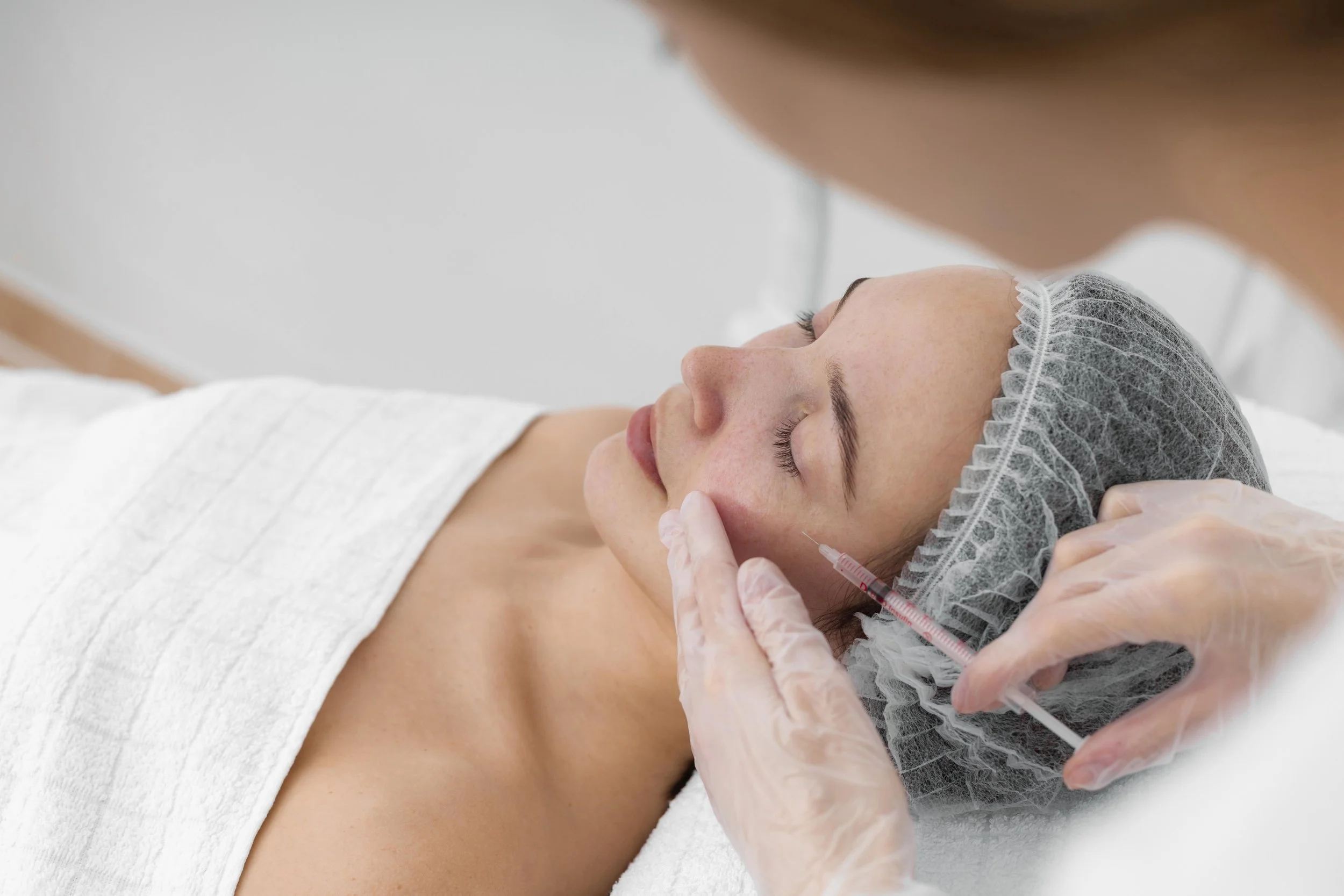 Person receiving a facial injection in a clinical setting, wearing a hair cap and covered with a white towel.