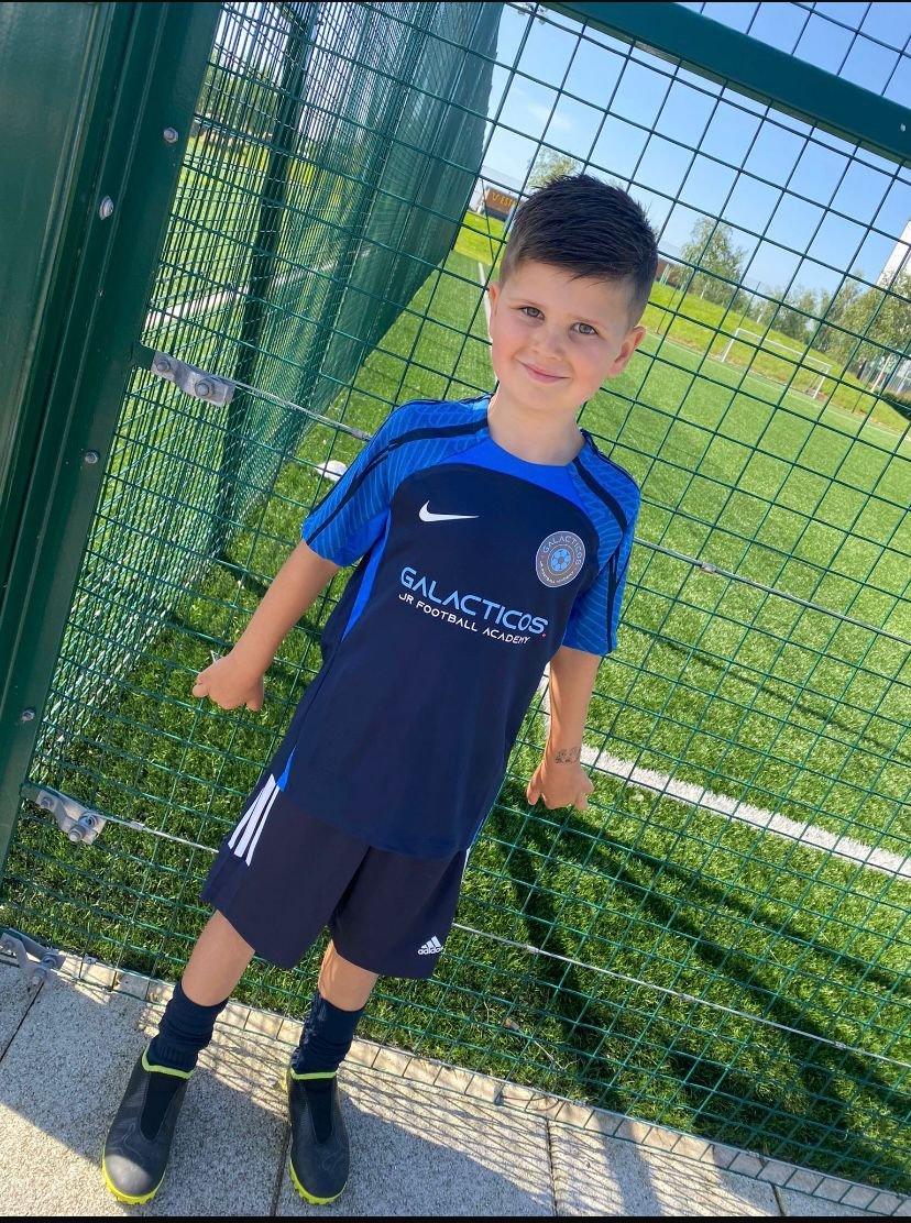 A young boy standing next to a green fence on a sunny day, wearing a blue and black soccer jersey, black shorts, black socks, and sports shoes, smiling at the camera. At Galacticos Junior Football Academy at York LNER Community Stadium.