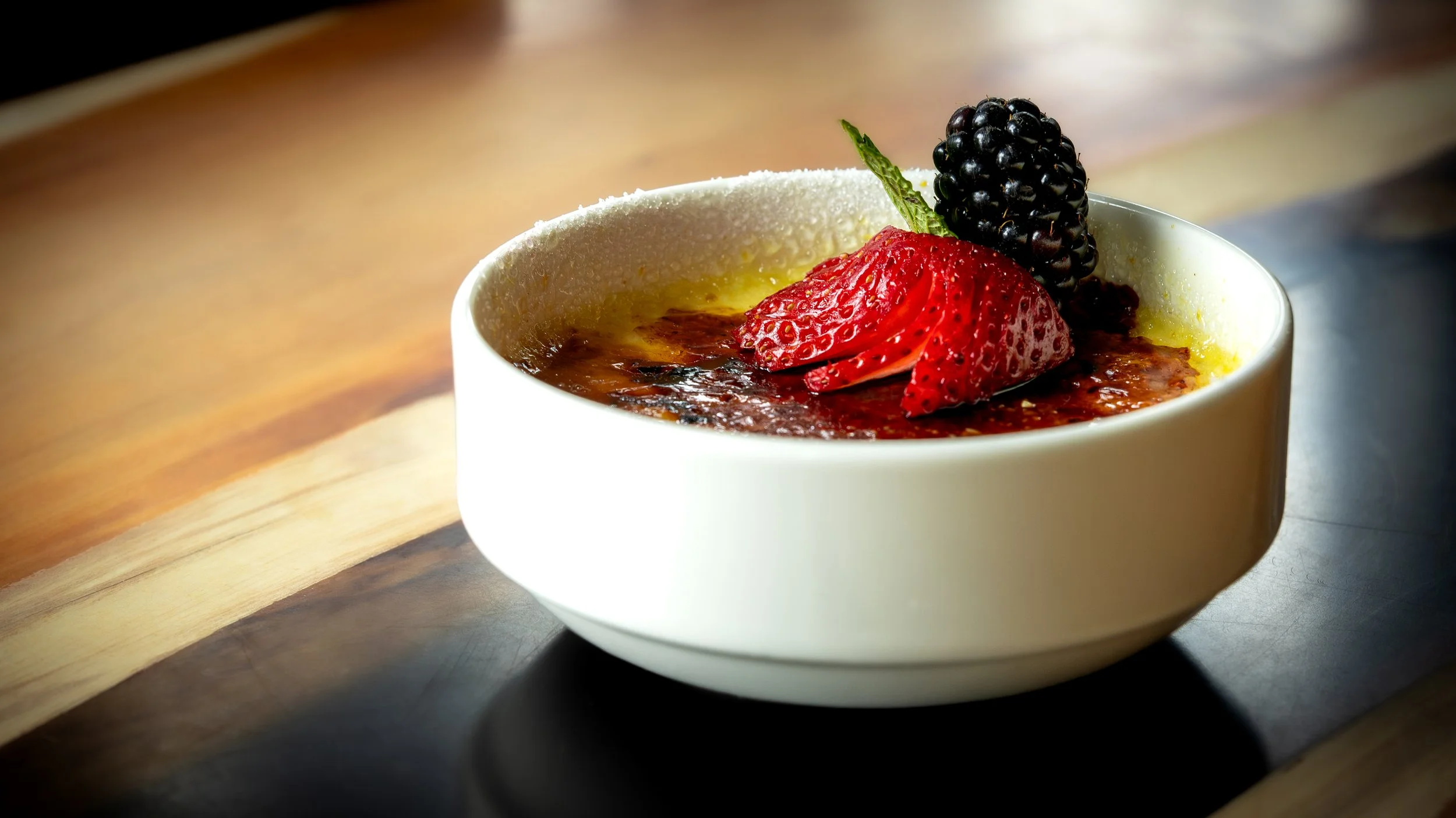 A white ceramic bowl filled with crème brûlée topped with strawberry slices, blackberry, and a green mint leaf on a wooden surface.