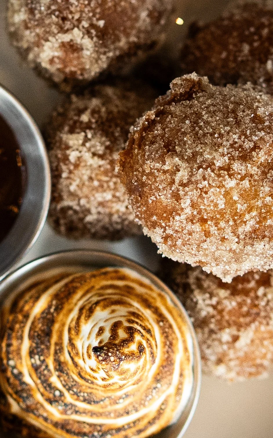 Close-up of a plate of sugar-coated balls, possibly donuts or fried desserts, along with a cup of coffee and other pastries in the background.