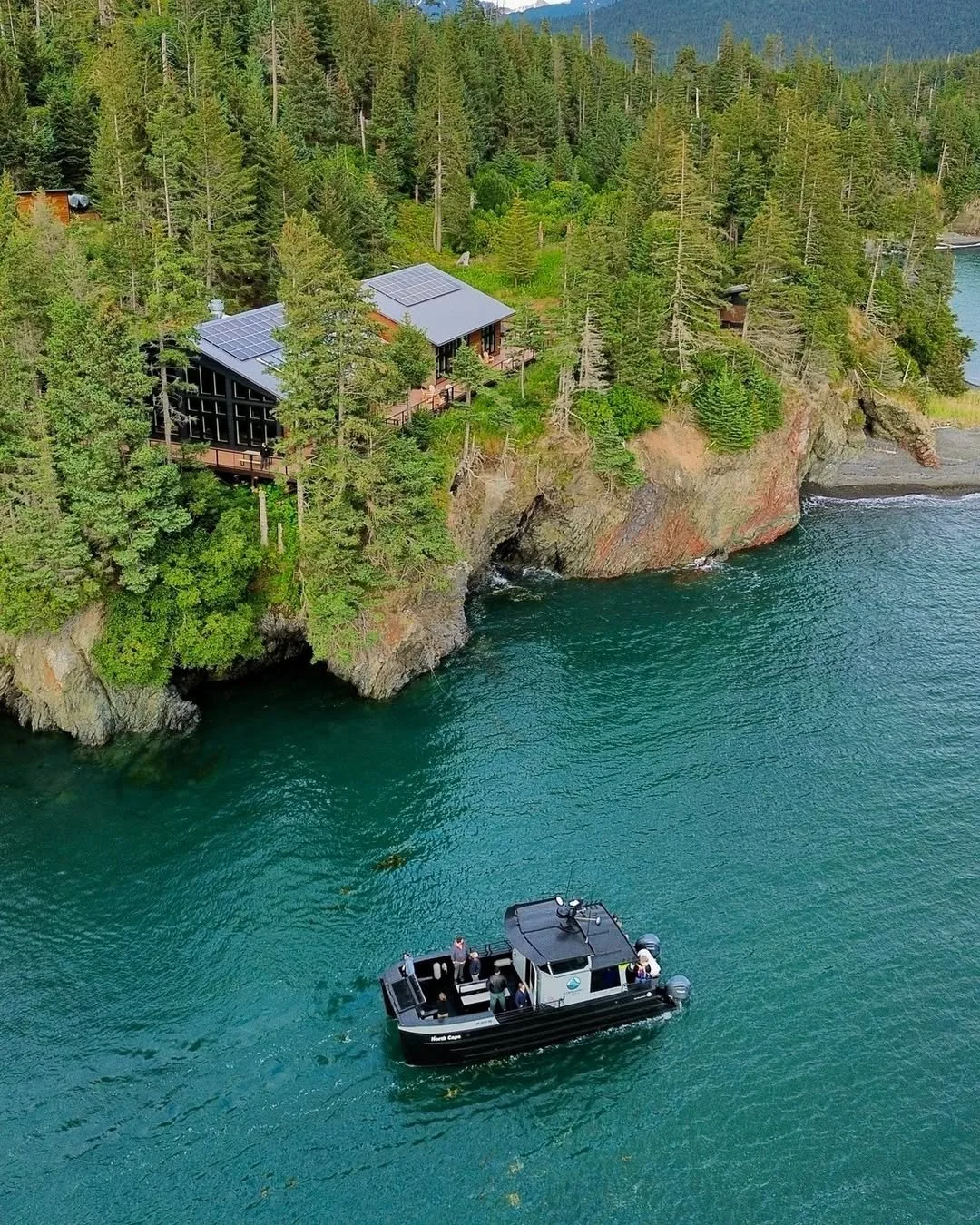 A house with solar panels situated on a forested cliff overlooking a body of water, with a black boat and several people in life jackets nearby.