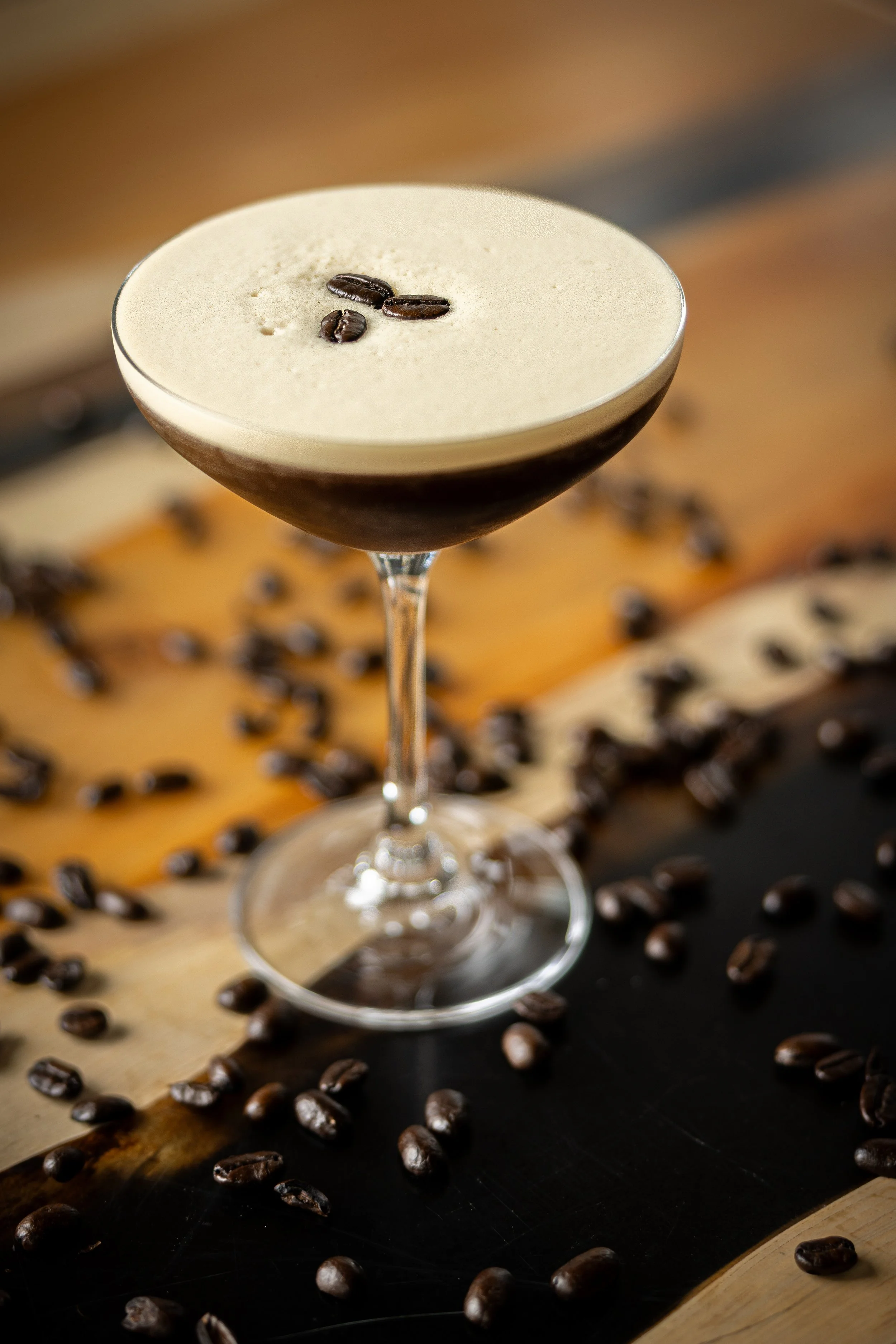 A coffee-based cocktail in a stemmed glass garnished with coffee beans, surrounded by scattered coffee beans on a wooden surface with a dark background.